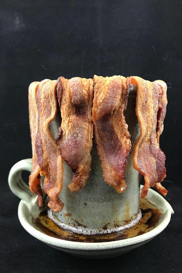 Pale green rustic mug-shaped cooker with cooked bacon draped on the rim and bacon grease in the tray at the bottom