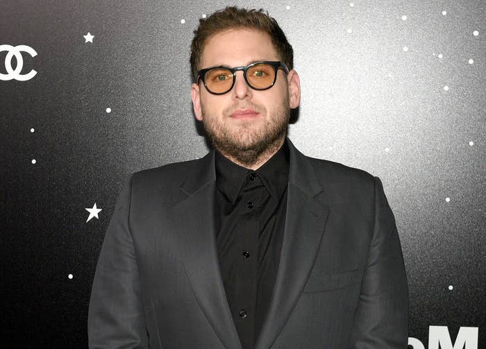 Jonah wears a black suit jacket and black button down with orange tinted glasses