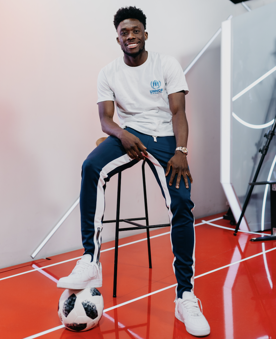 Alphonso Davies sitting on a stool with his foot on a soccer ball.