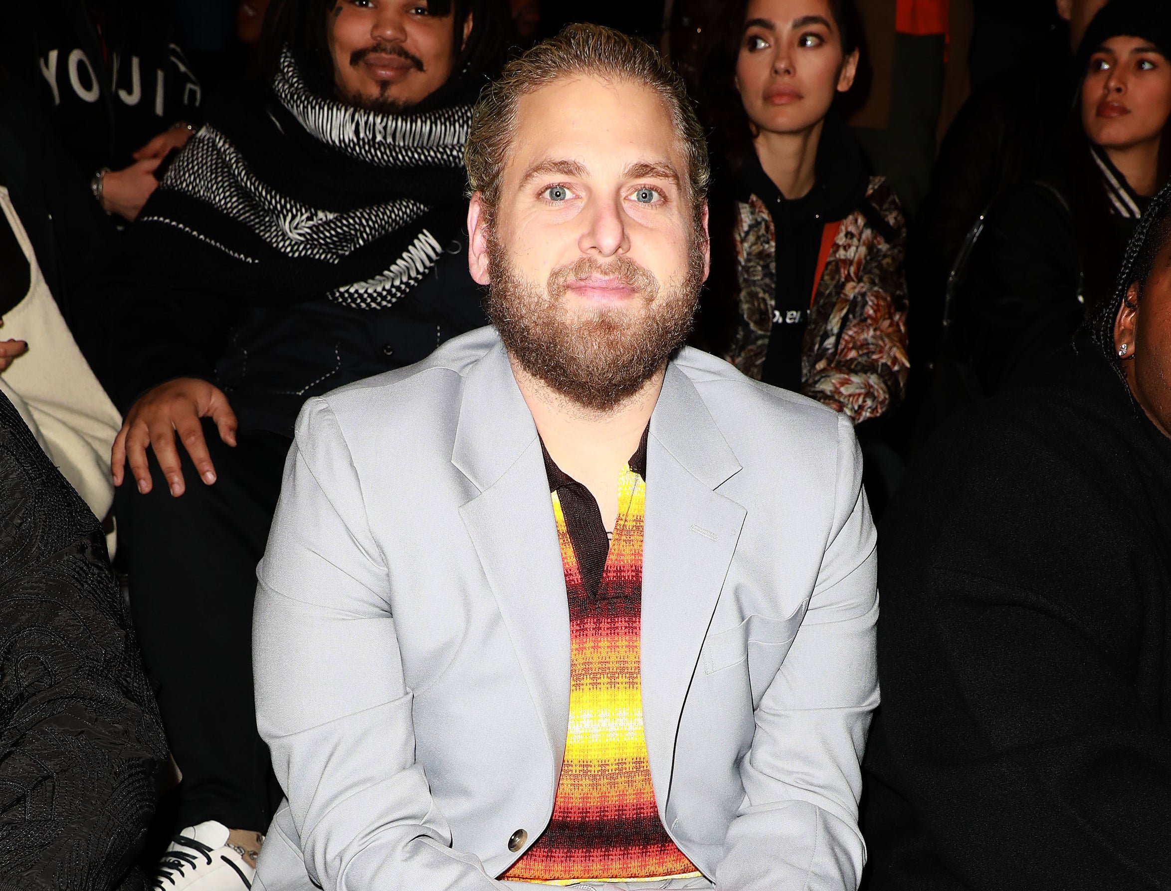 Jonah wears a grey suit with a yellow and orange knit polo underneath