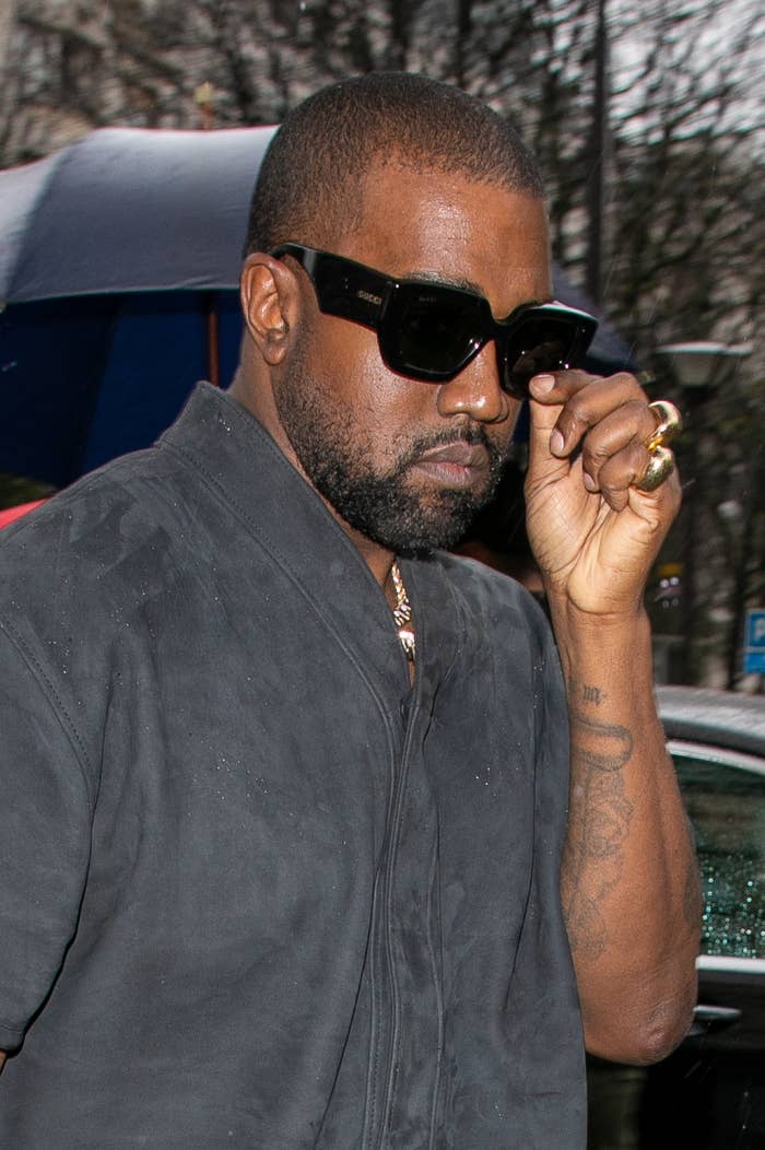West puts his hand in front of his face while wearing sunglasses and a button-down shirt