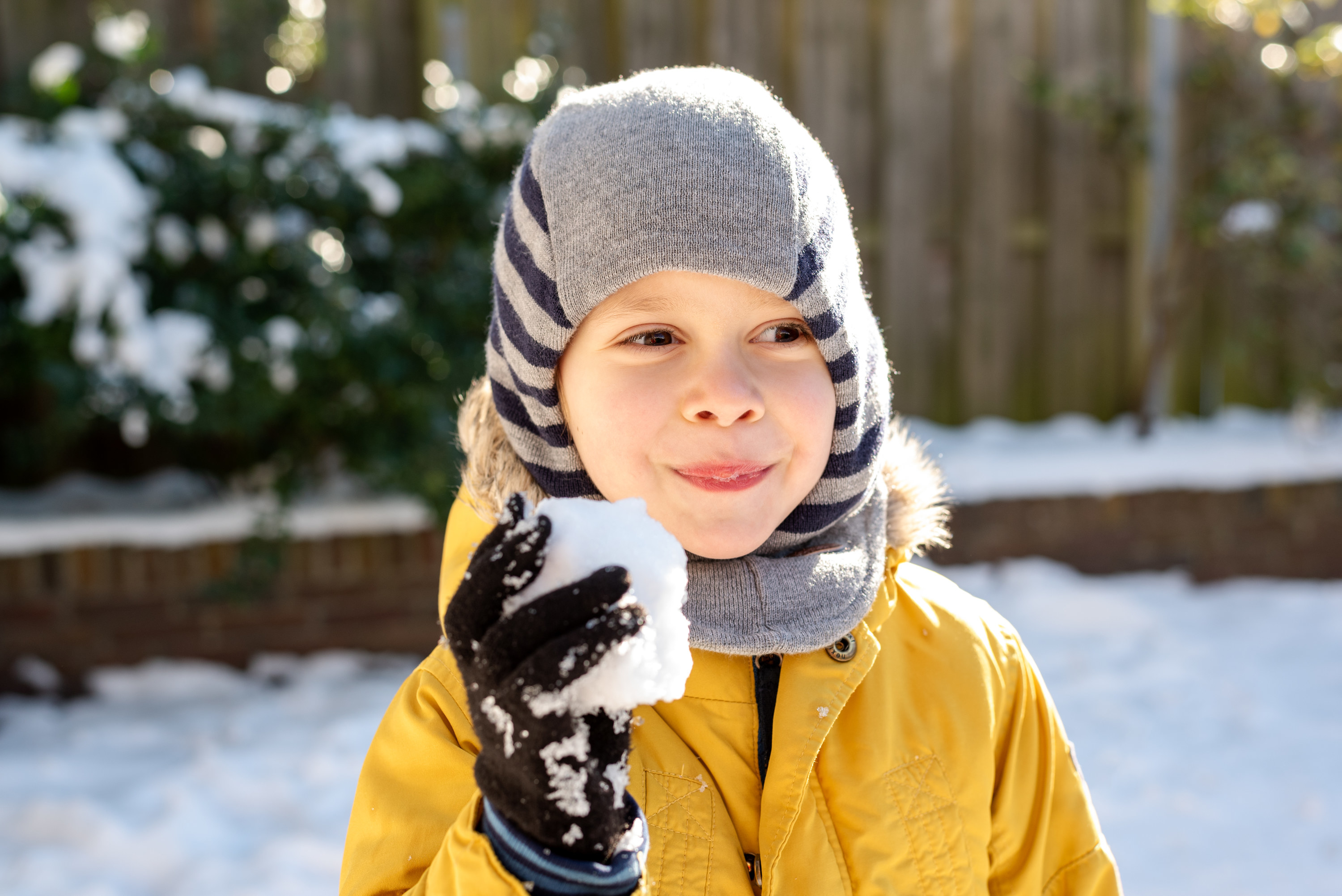 A kid outside in coat and warm hat smiling, holding a snowball in gloved hand