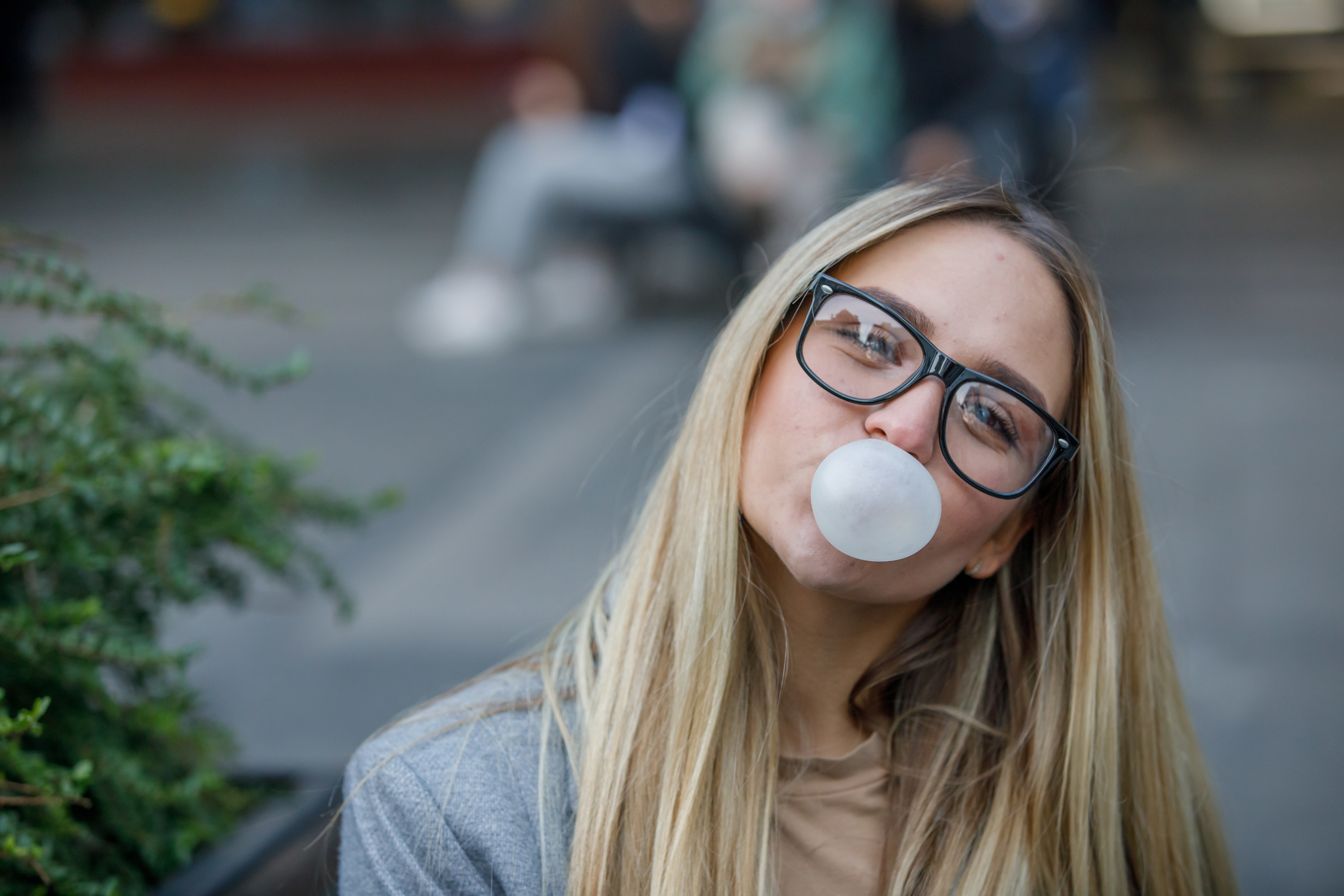 A young woman with long hair and glasses blowing a big bubble with gum