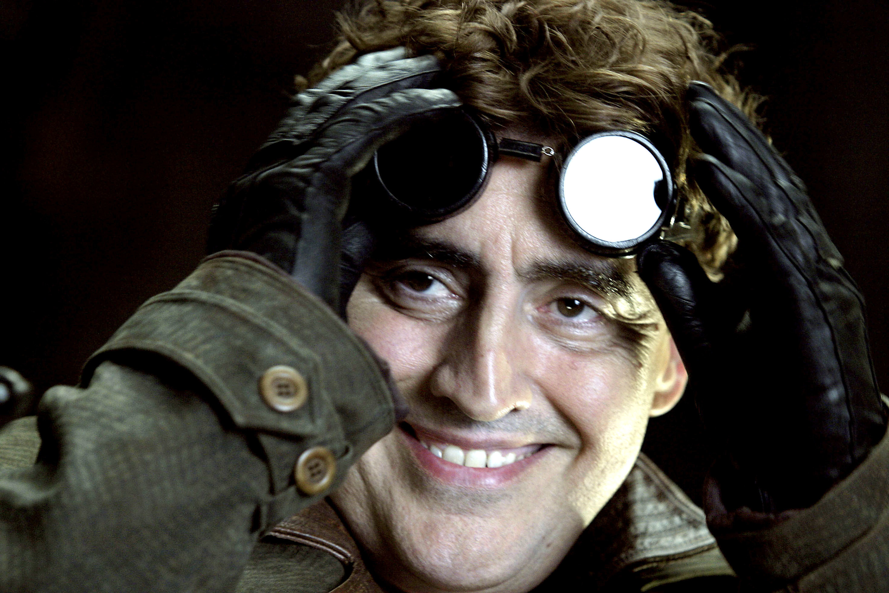 Alfred Molina smiles as he pushes his goggles off of his forehead and pushes up his shaggy hair as Doc Ock in Spider-Man 2