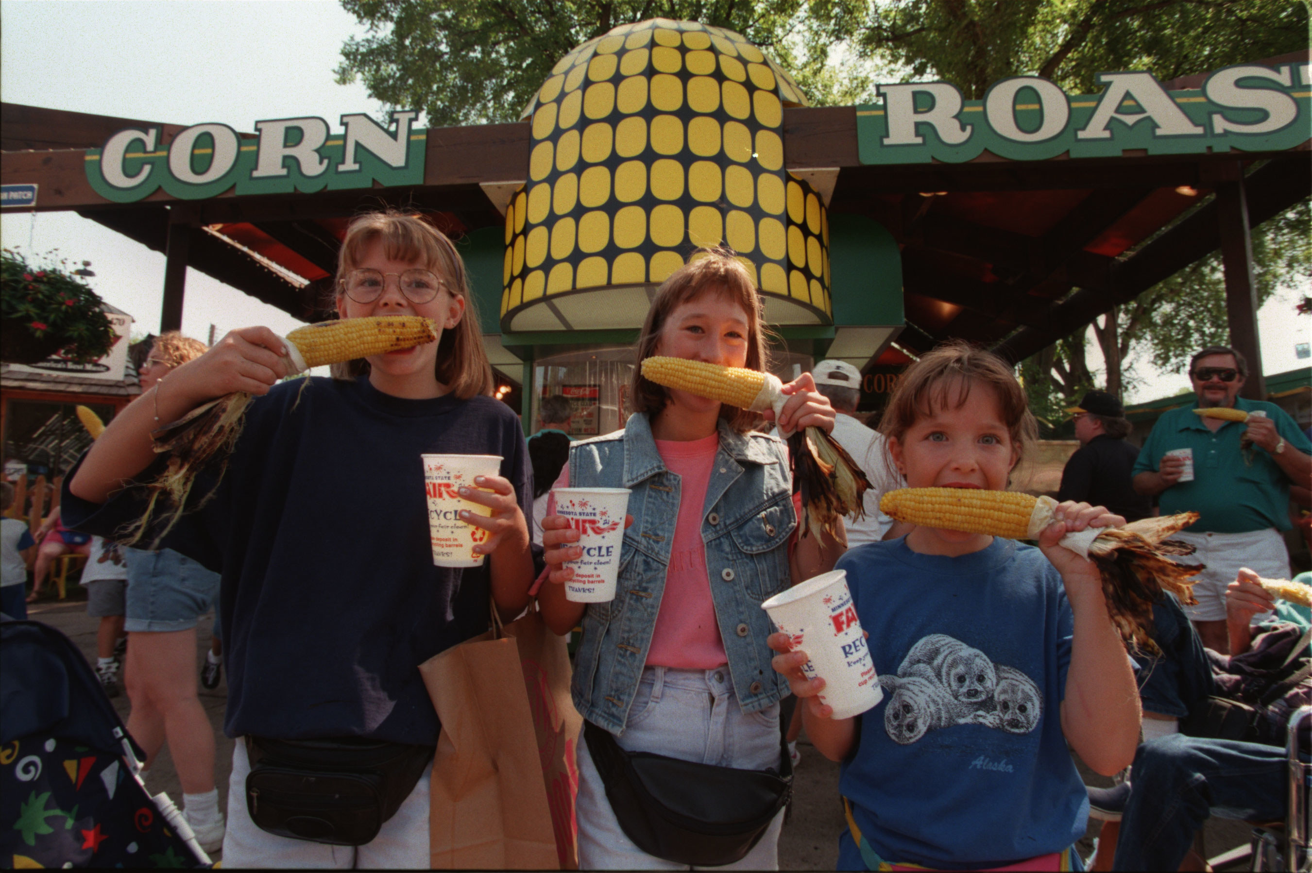 Three young females hold up corn on the cob and hold cups at the state fair