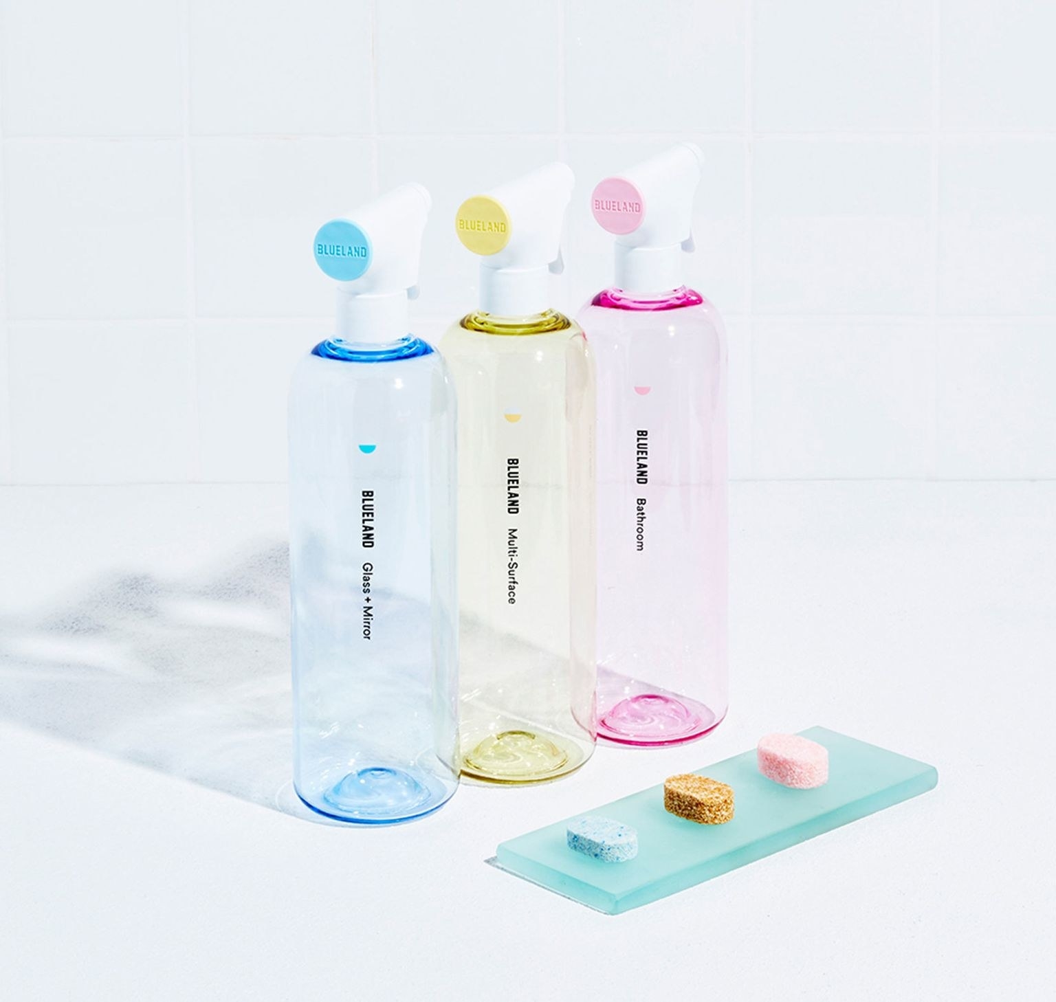 three plastic bottles — one blue, one yellow, and one pink — next to three dissolvable cleaning tablets