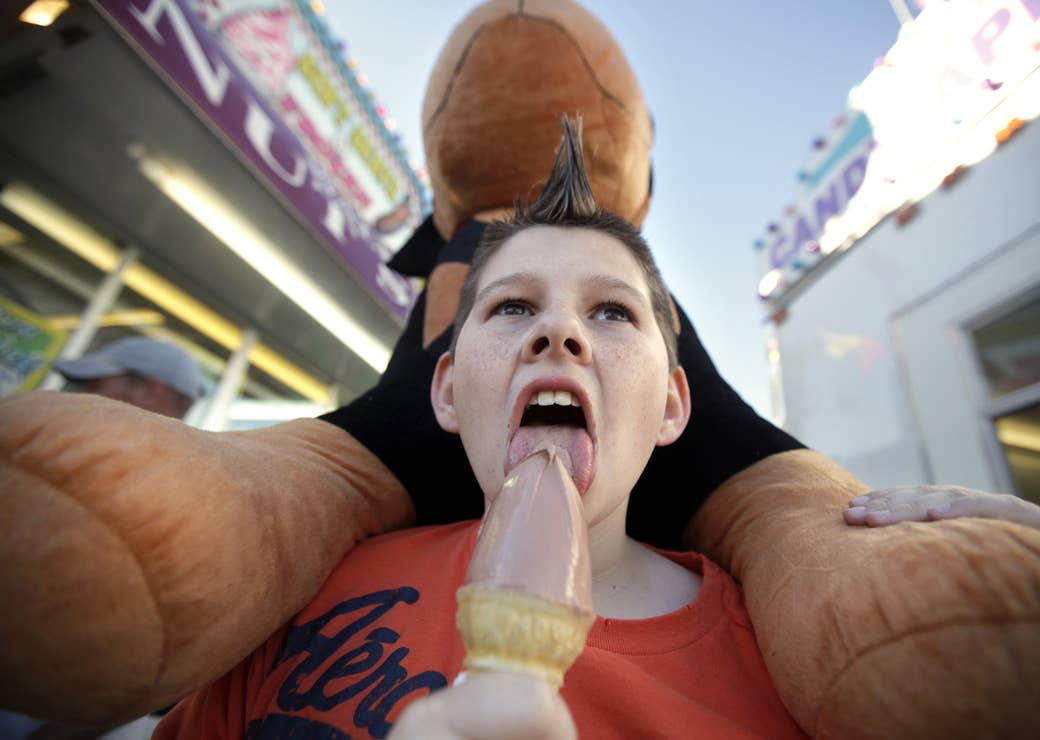 A child licks an ice cream cone and carries a stuffed dog on his shoulders while attending a fair