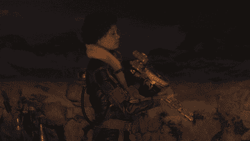 Val loads a gun while standing by a campfire