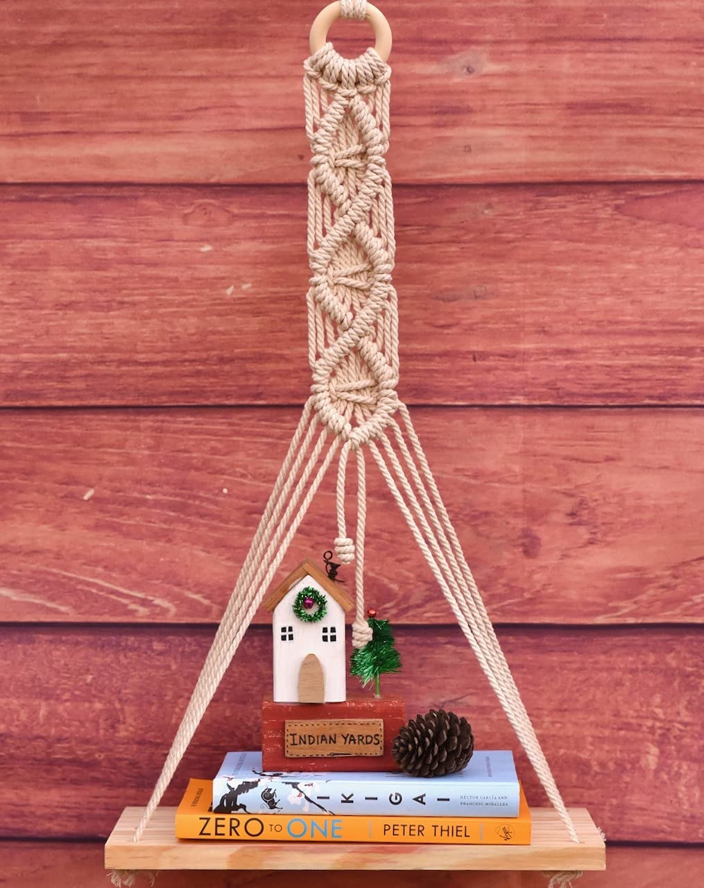 A macrame wall hanging with a wooden shelf holding a few books and showpieces