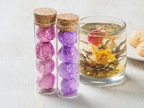 two containers with one filled with pink foil balls and the other with purple foil ball and a clear tea cup with tea and a blooming yellow flower inside