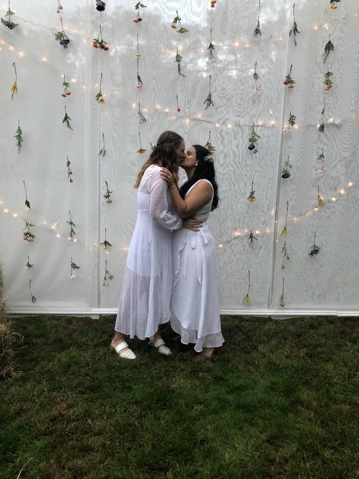 Marisa (left) and her wife (right) are kissing in front of a white backdrop. They are wearing wedding dresses, and strings of flowers and lights are behind them. 