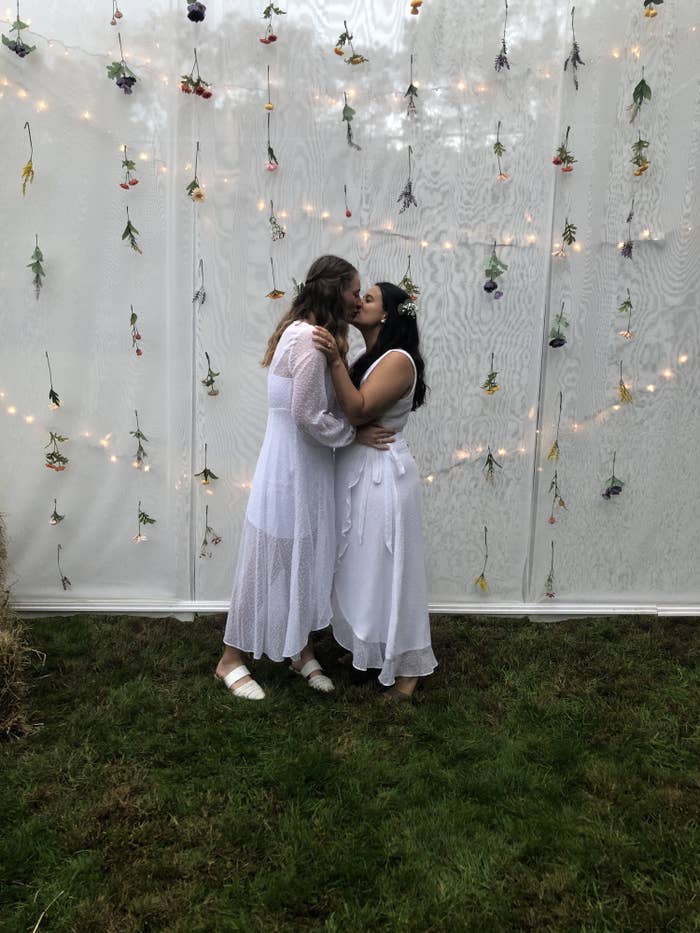 Marisa (left) and her wife (right) are kissing in front of a white backdrop. They are wearing wedding dresses, and strings of flowers and lights are behind them.