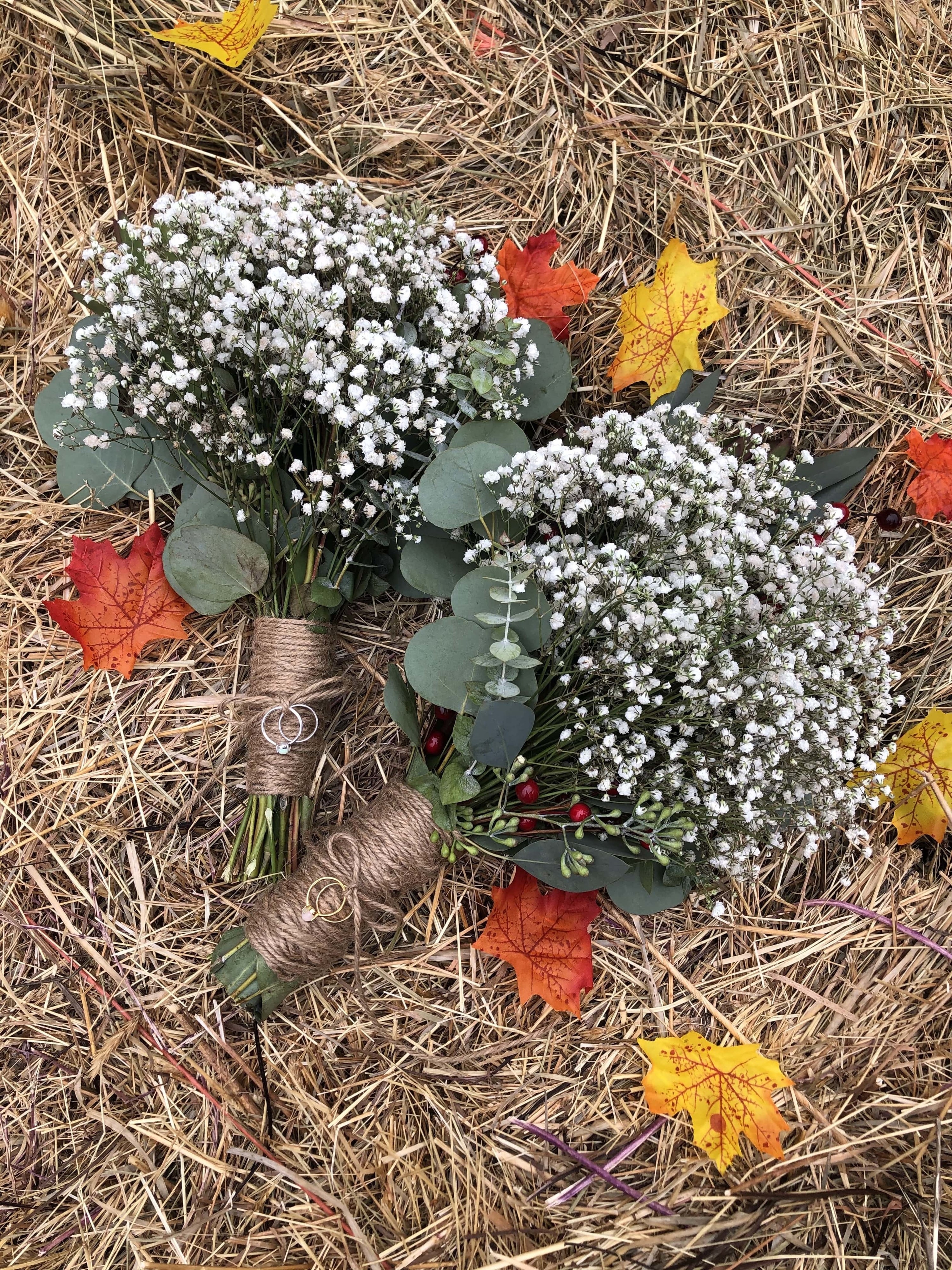 Two bouquets of baby&#x27;s breath and eucalyptus leaves. The stems of the flowers are wrapped in brown string. The top bouquet has a silver engagement ring and wedding band tied to it, and the bottom bouquet has gold rings. 