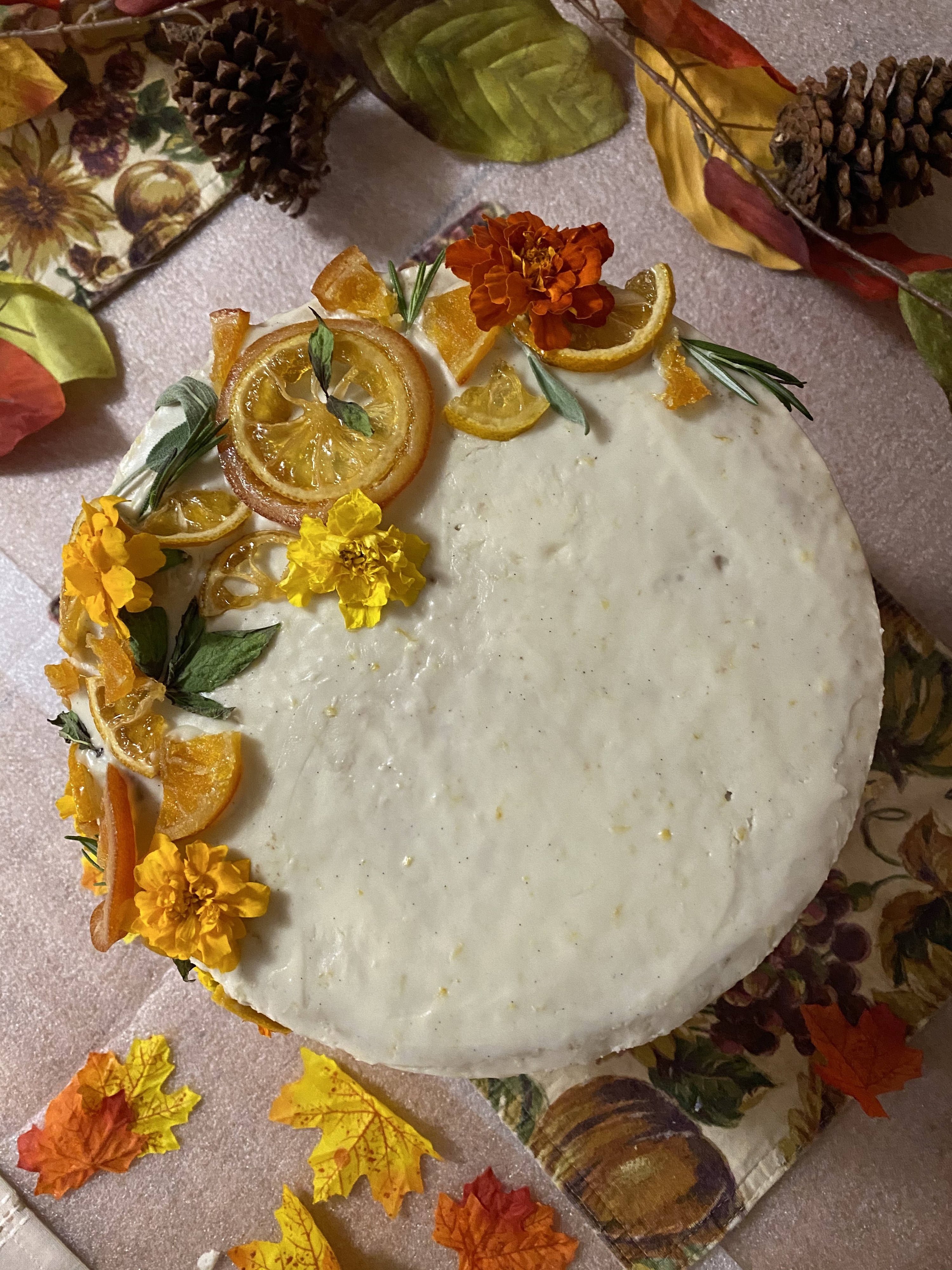 A white cake, decorated with yellow edible flowers, orange and lemon slices, and green herbs. 