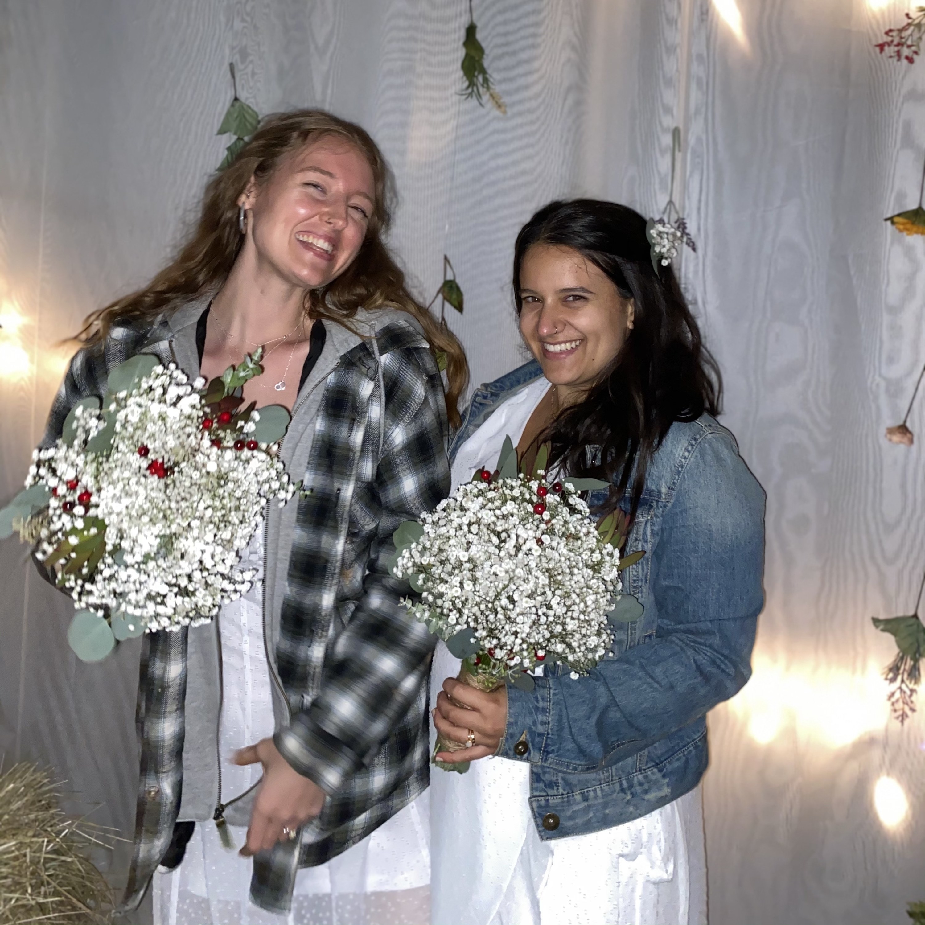 Marisa (left) and her wife (right) are grinning at the camera. They have jackets on over their wedding dresses, and are holding their bouquets. 