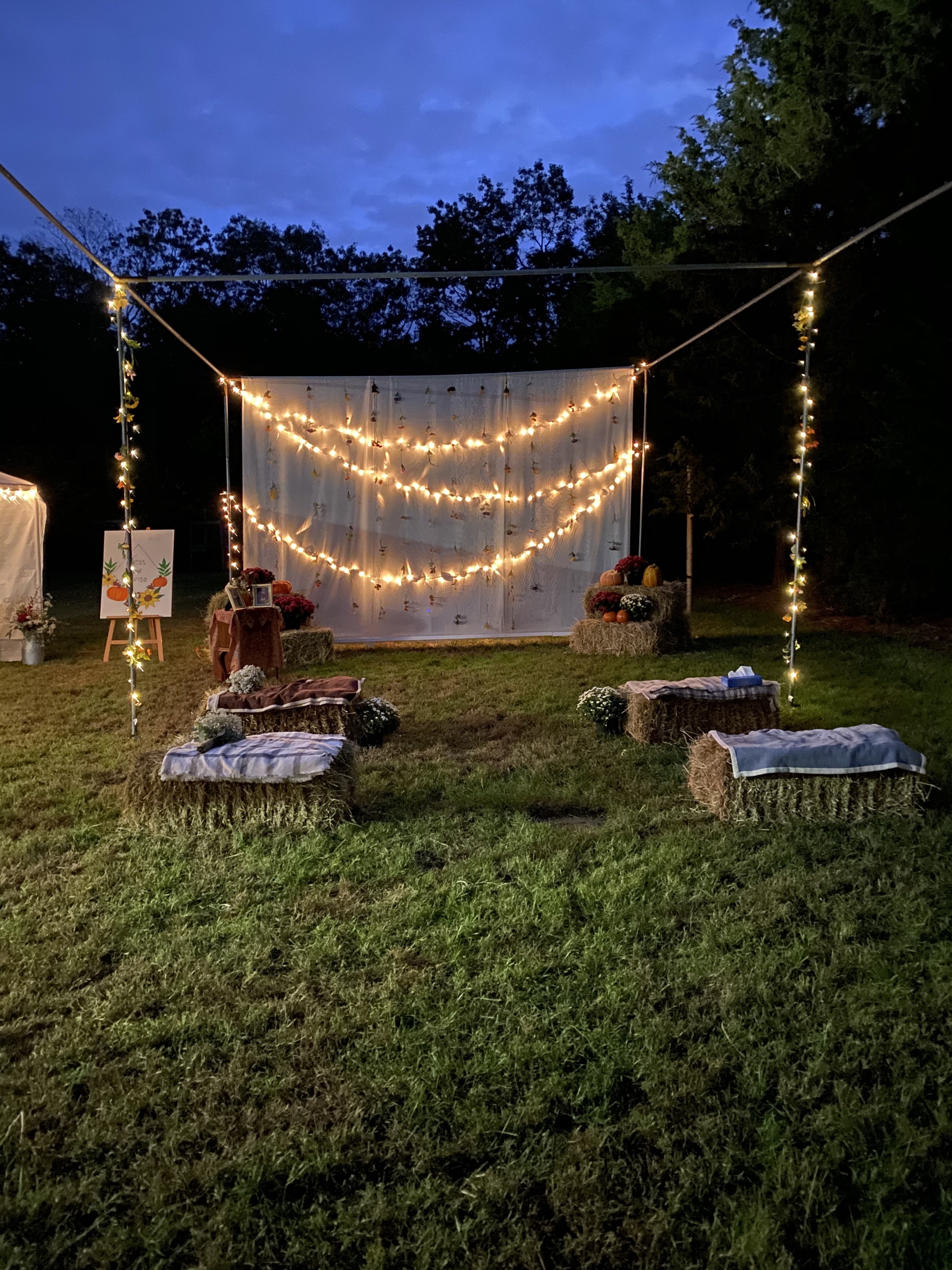 The frame of a batting cage is filled with hay bales with blankets, and decorated with string lights and a white backdrop. 