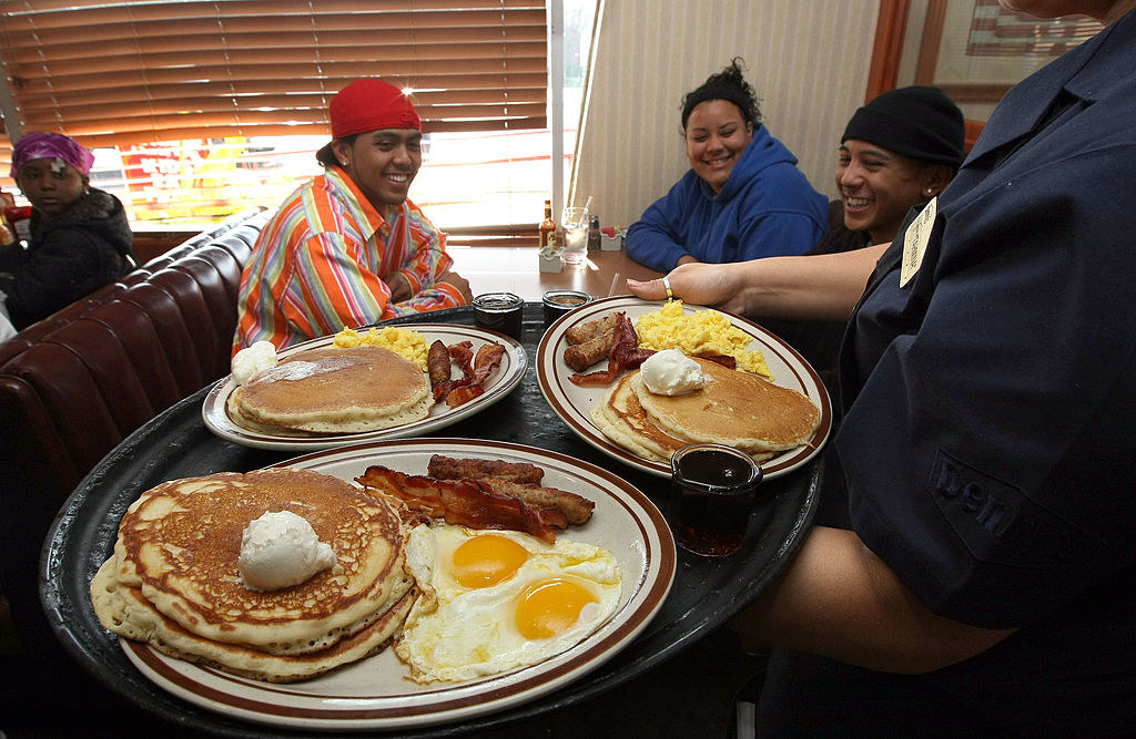 Three smiling people sitting at a table in a diner looking at plates of pancakes, sausage/bacon, and eggs that they're about to be served