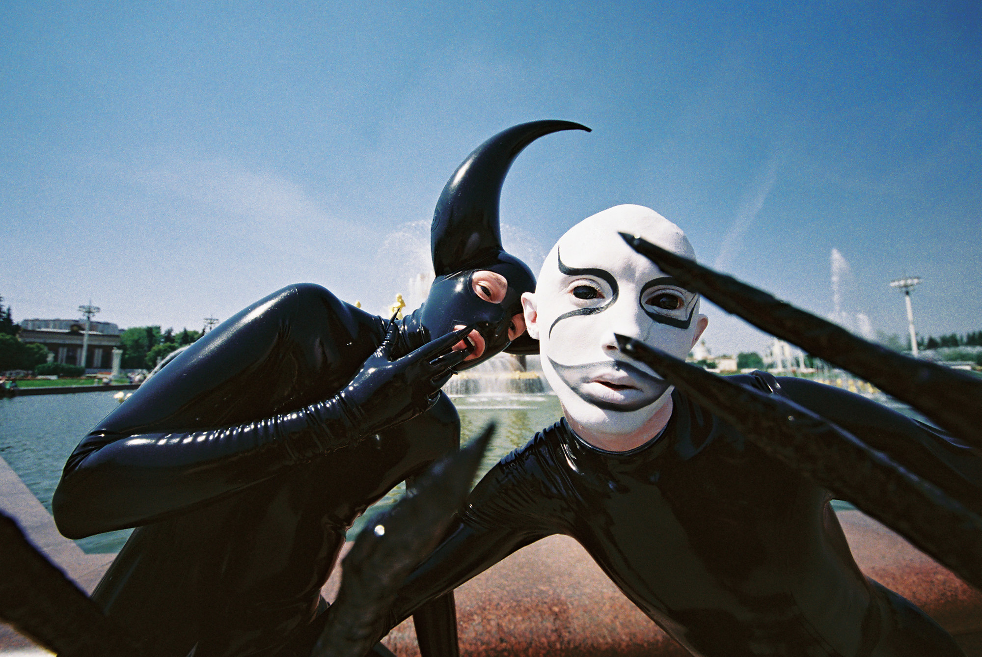 Two people in latex suits in front of a fountain, one with face paint and one with a mask