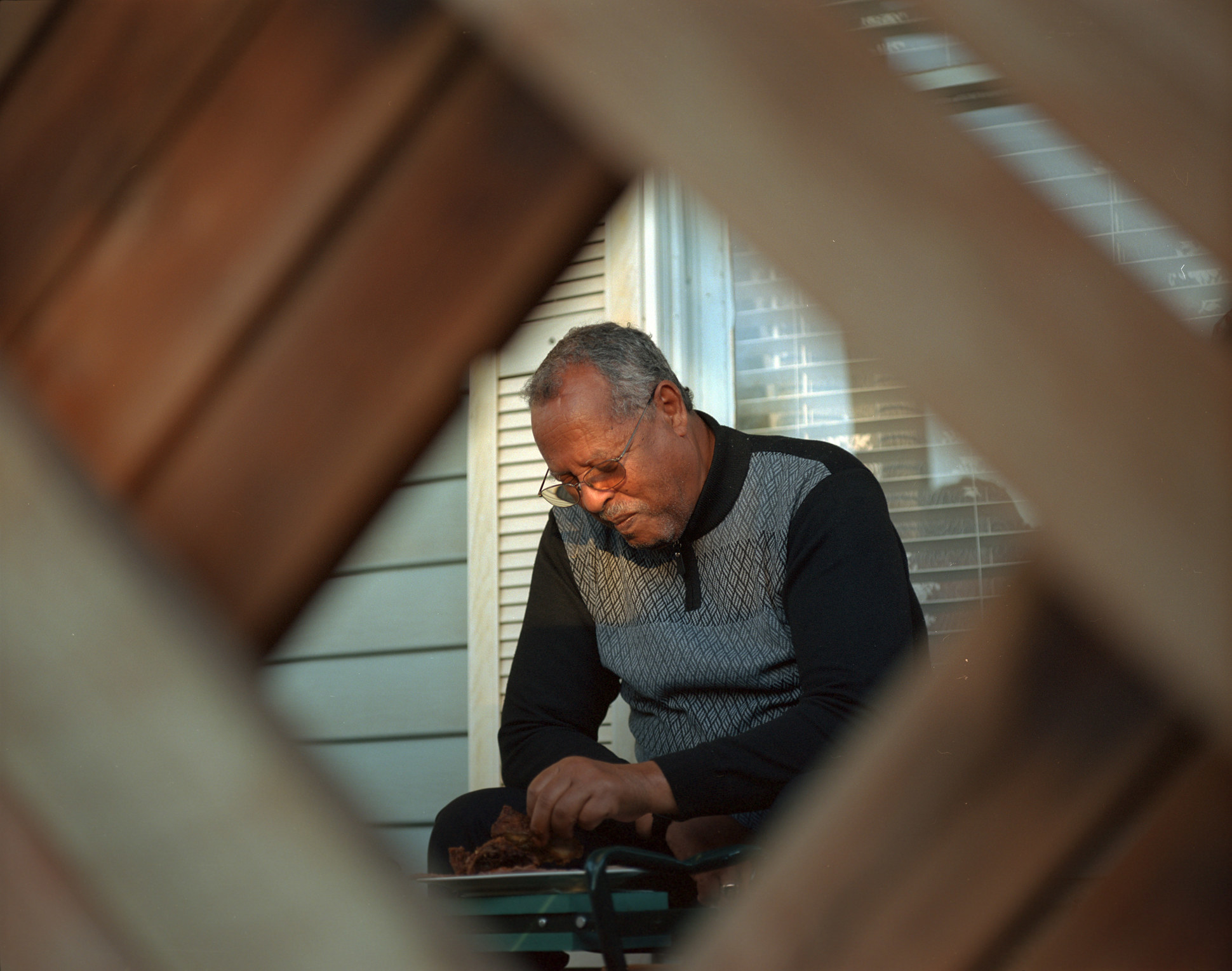 The photographer&#x27;s father eating in the garden, shot through a fence