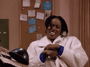 Maxine Shaw holding her white coat closed while she maniacally laughs and flaps her whiite gloves. 