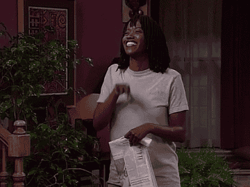 Erika Alexander as Maxine Shaw smiling and waving while holding a newspaper.