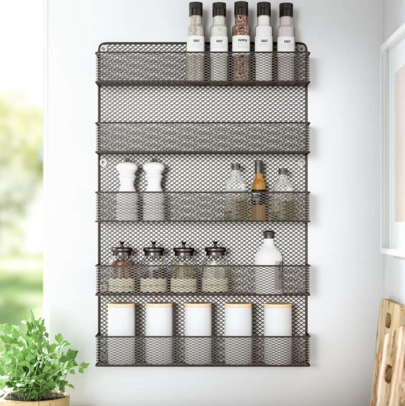 The spice rack is mounted on a white wall and has five rows holding various sizes bottles, and it&#x27;s all against some greenery in the background
