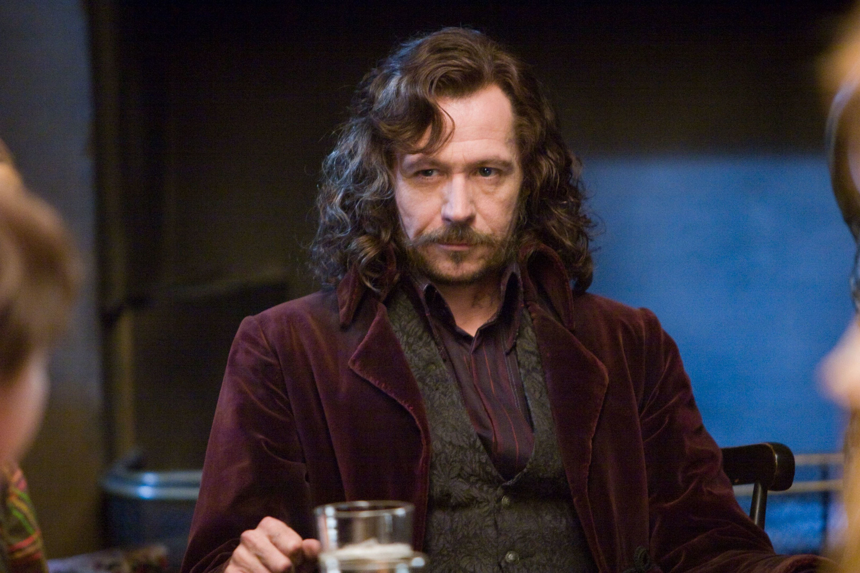 Gary Oldman sits at a table while looking at another character with a glass in front of him