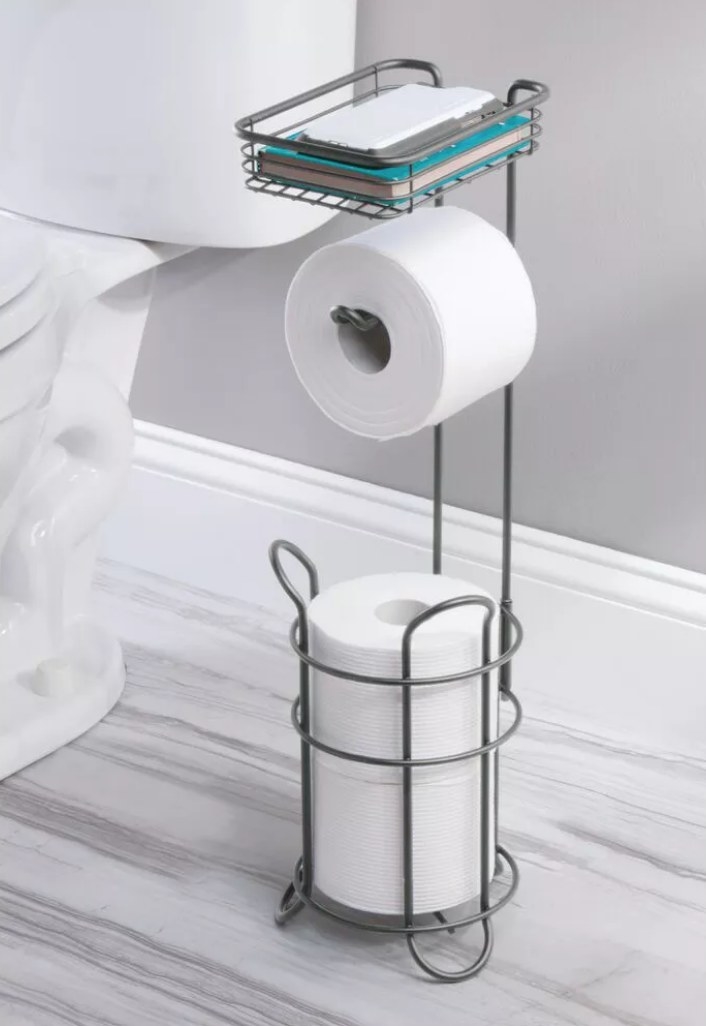 A graphite, metal freestanding toilet paper holder with a shelf atop filled with a phone and a book