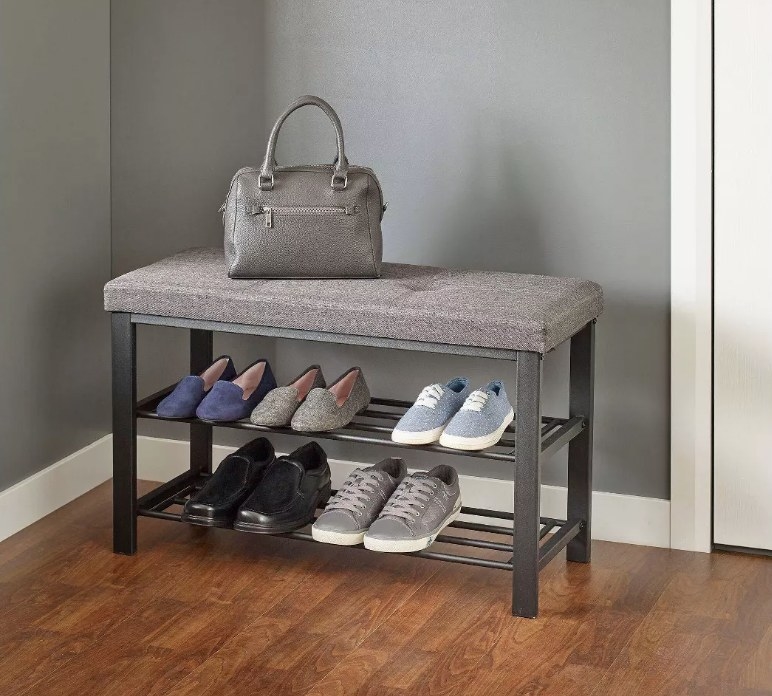 A grey, upholstered bench with two shelves below filled with shoes