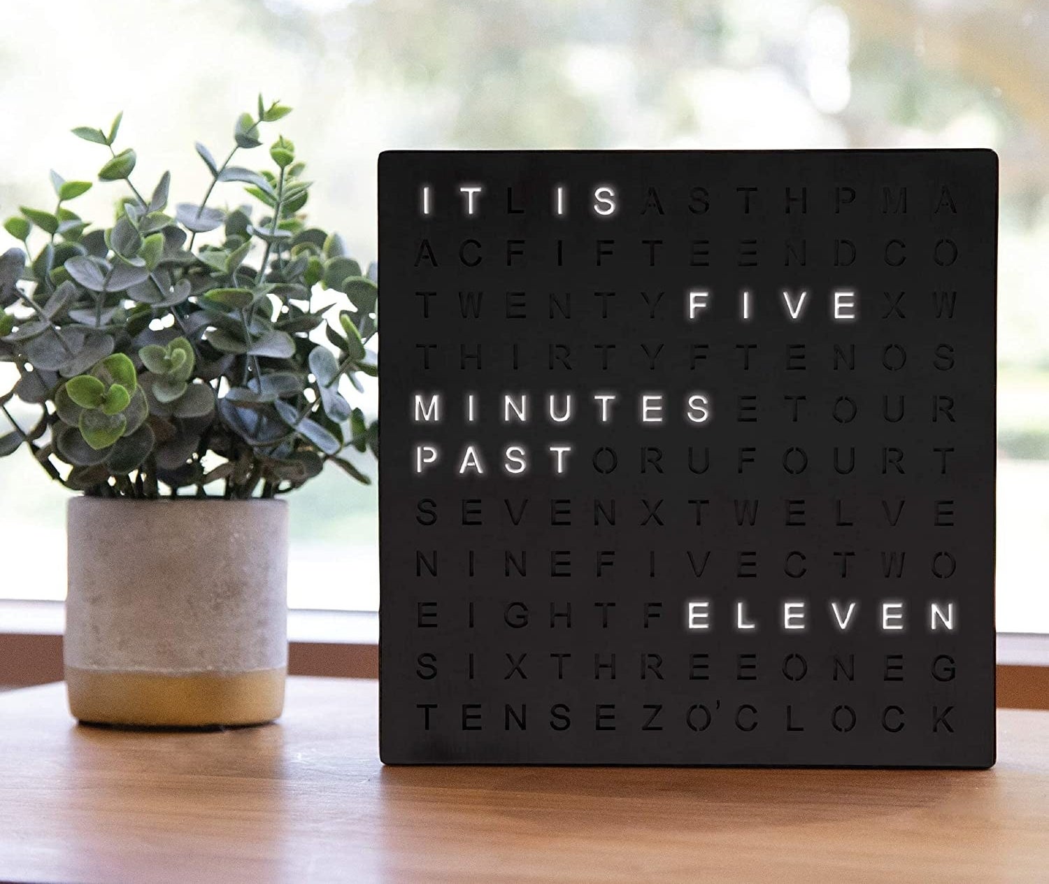 the word clock next to potted plant reading "It is five minutes past eleven"
