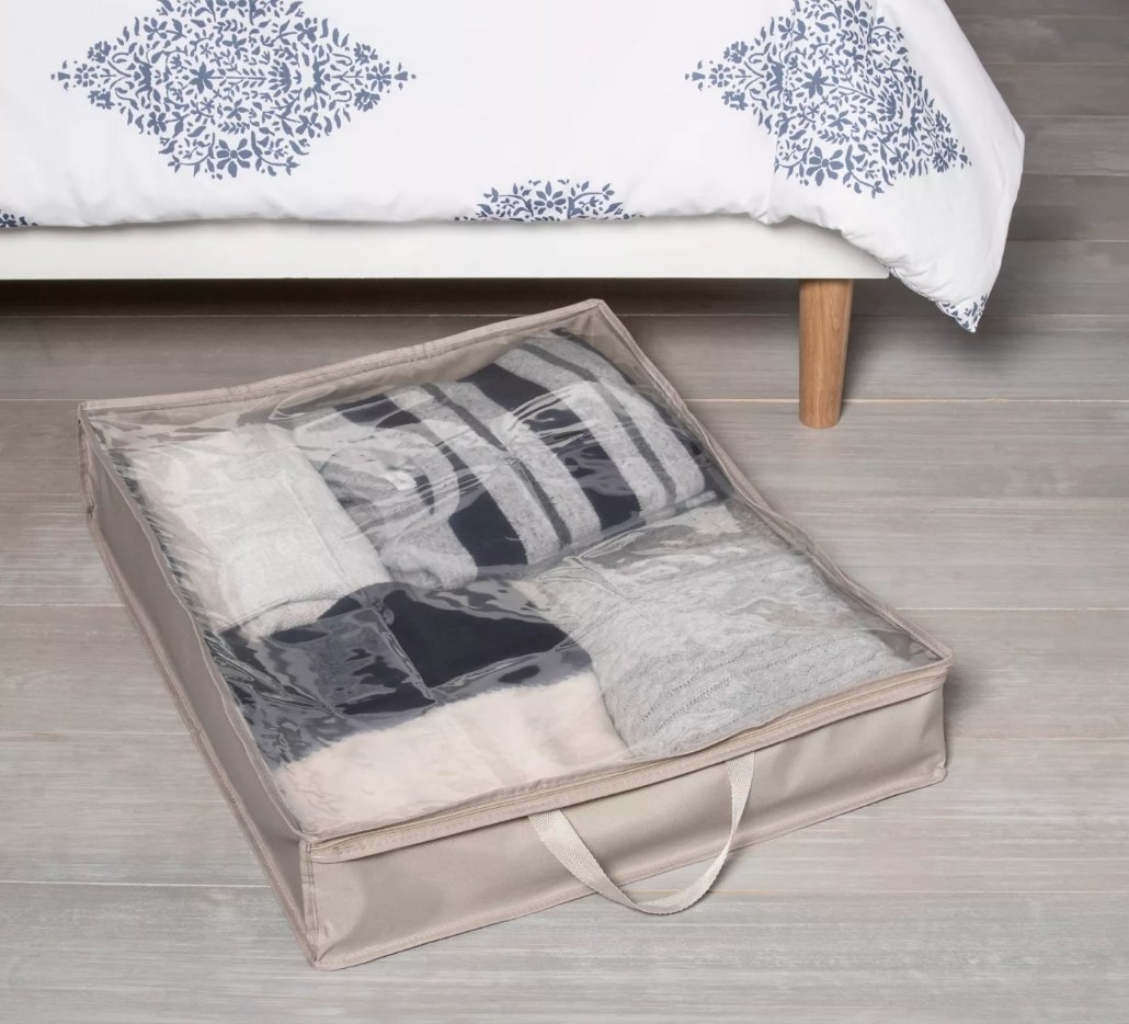 A fabric, zippered storage organizer filled with clothing and bedding under a bed