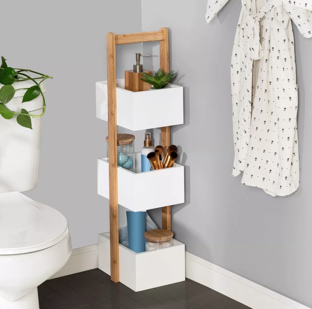 A bamboo, 3-tier bathroom storage caddy with three white caddies filled with makeup, soaps, brushes, etc