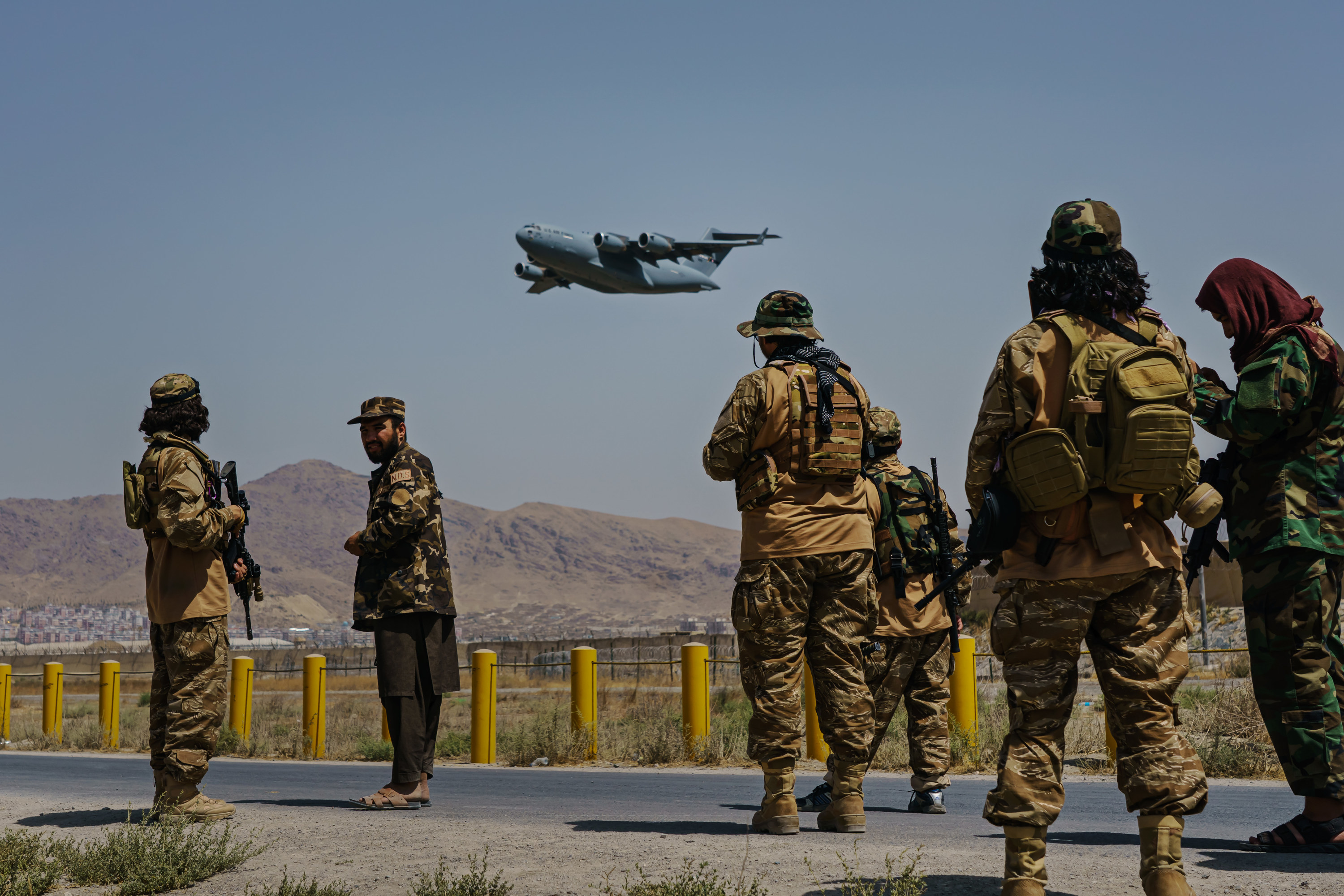 Taliban fighters stand outside Hamid Karzai International Airport as a plane flies over