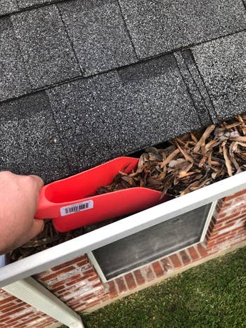 hand uses red gutter scoop to scoop up leaves from roof gutter