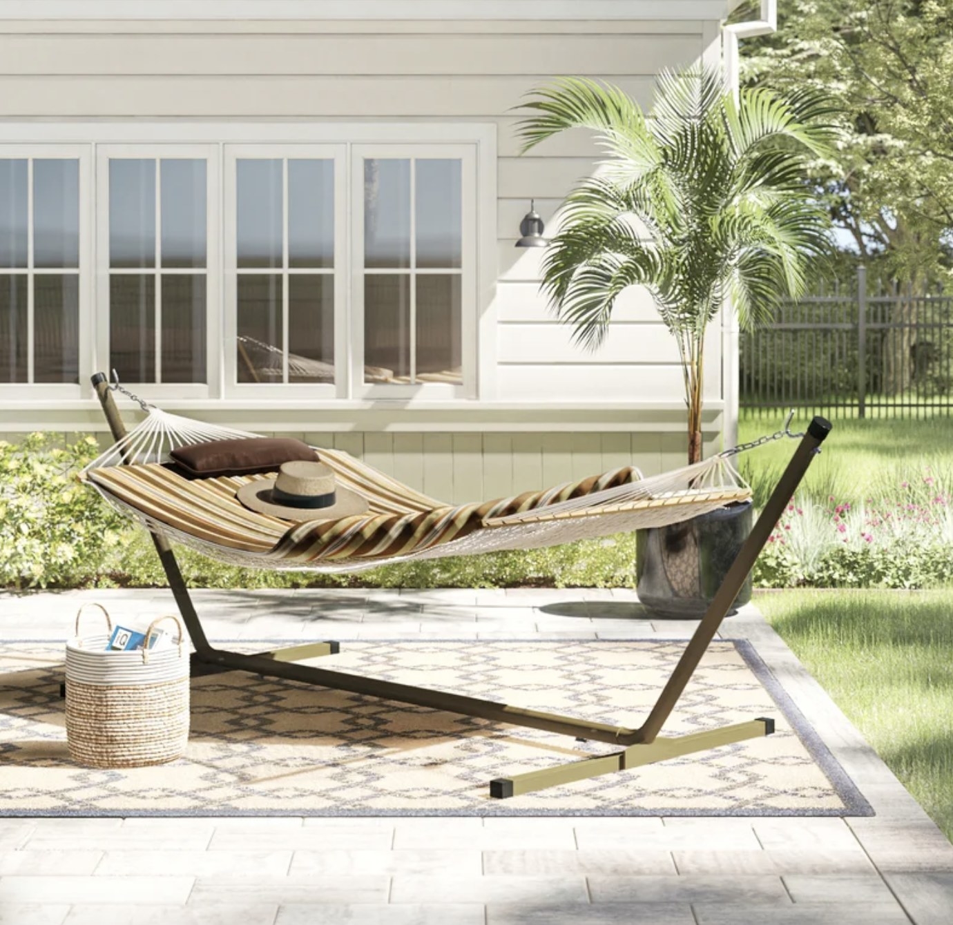 The white woven hammock has a green and tan stripe cover and brown pillow, and it&#x27;s set up outside in the sunshine