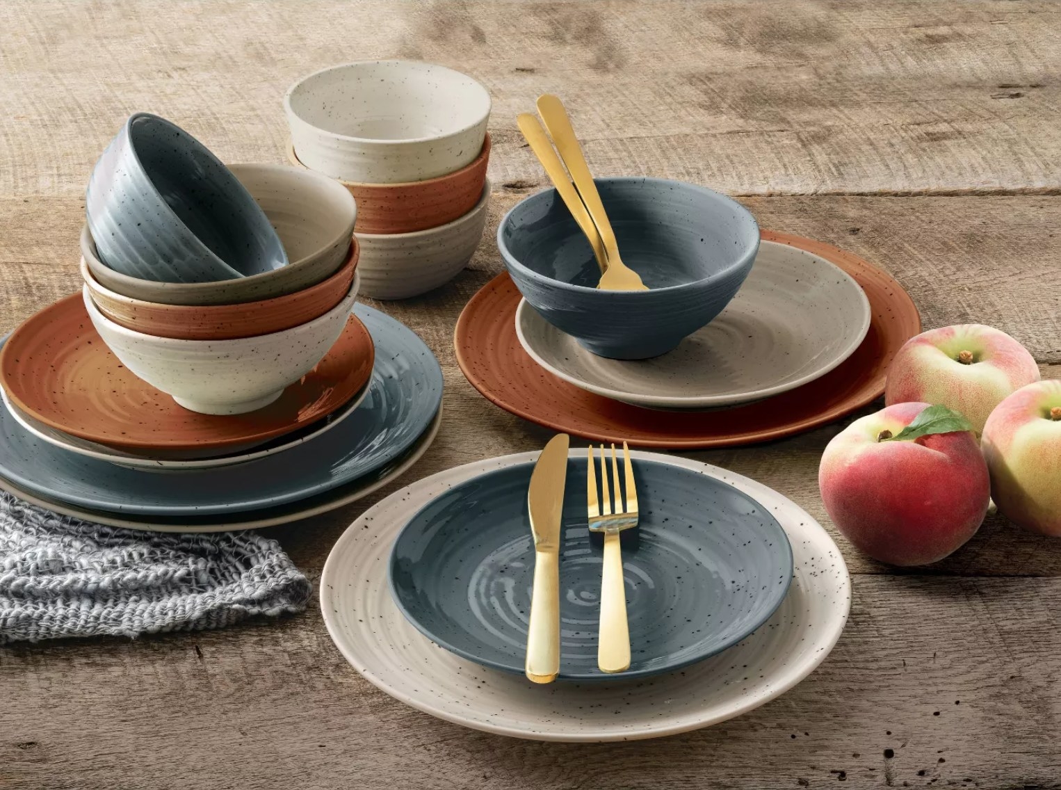 the full set of dinnerware in different colors, cream, blue, orange, and tan all stacked together with gold knives and forks on top and three apples on the side