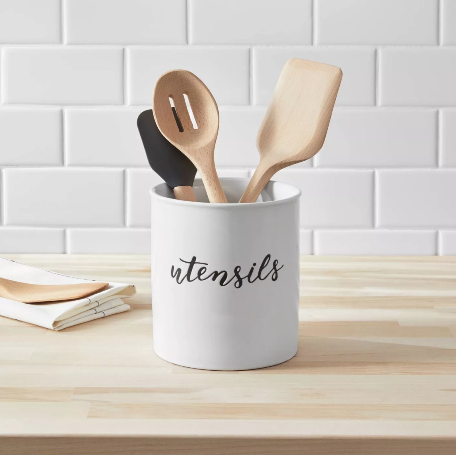 the white holder with &quot;utensils&quot; written in black cursive on a wooden counter with a slotted turner, spatula, and scraper inside