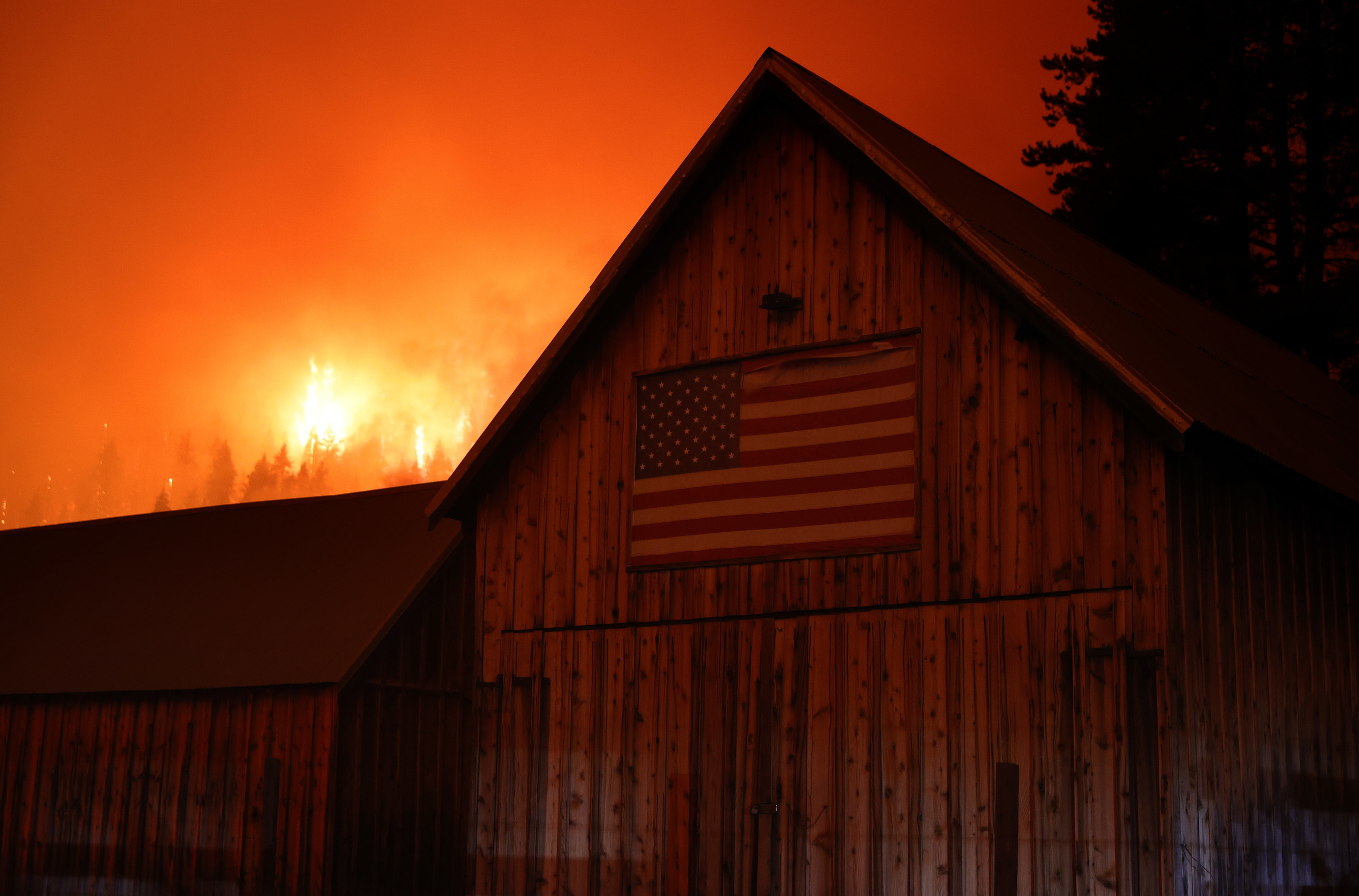 A fire blazes on a hillside in the background behind a barn with a US flag on the side