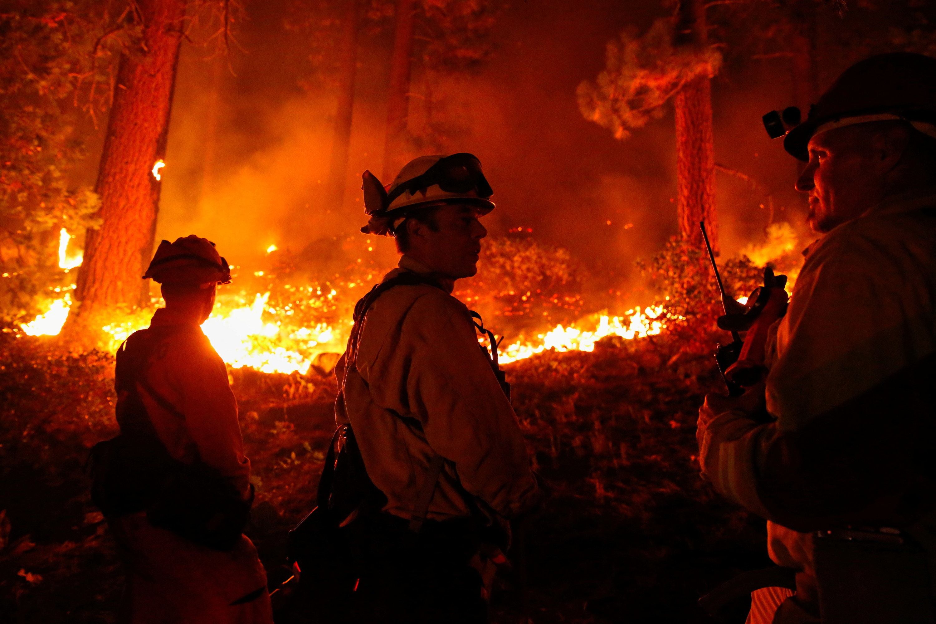 Three firefighters in uniform stand near a brush fire and converse