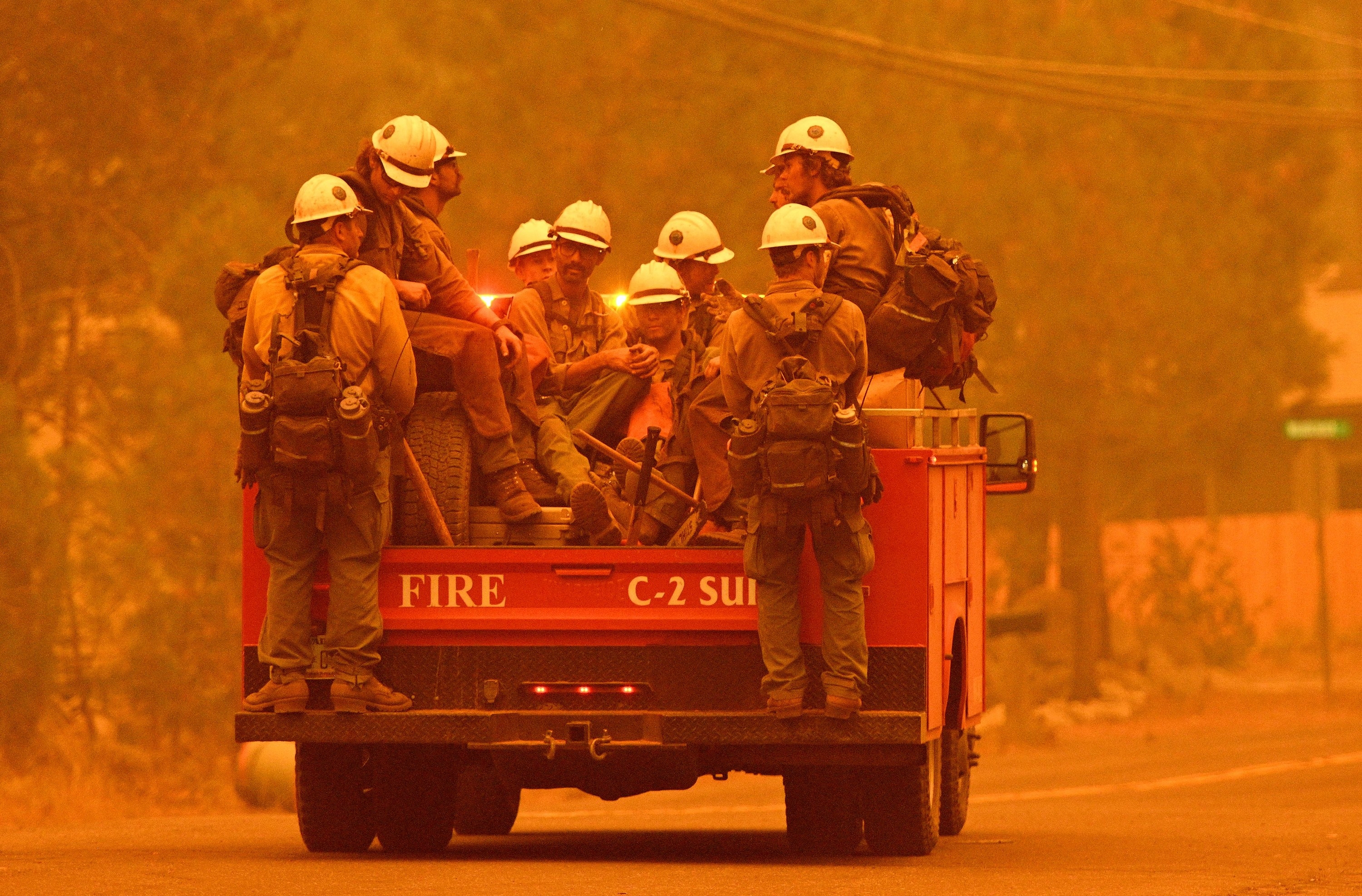 Ten firefighters in uniform sit and stand on a moving truck under a hazy sky