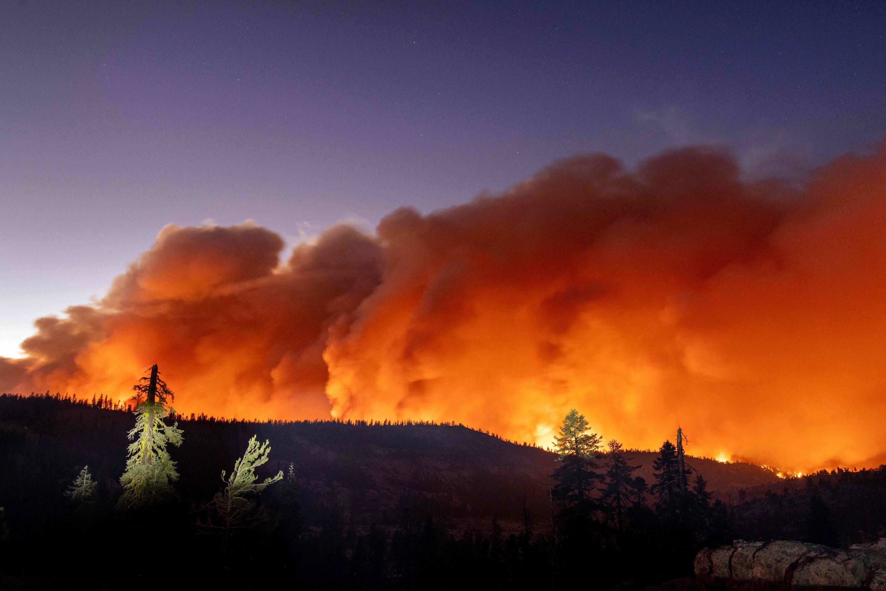 A massive cloud of smoke fills the sky over a forested ridge