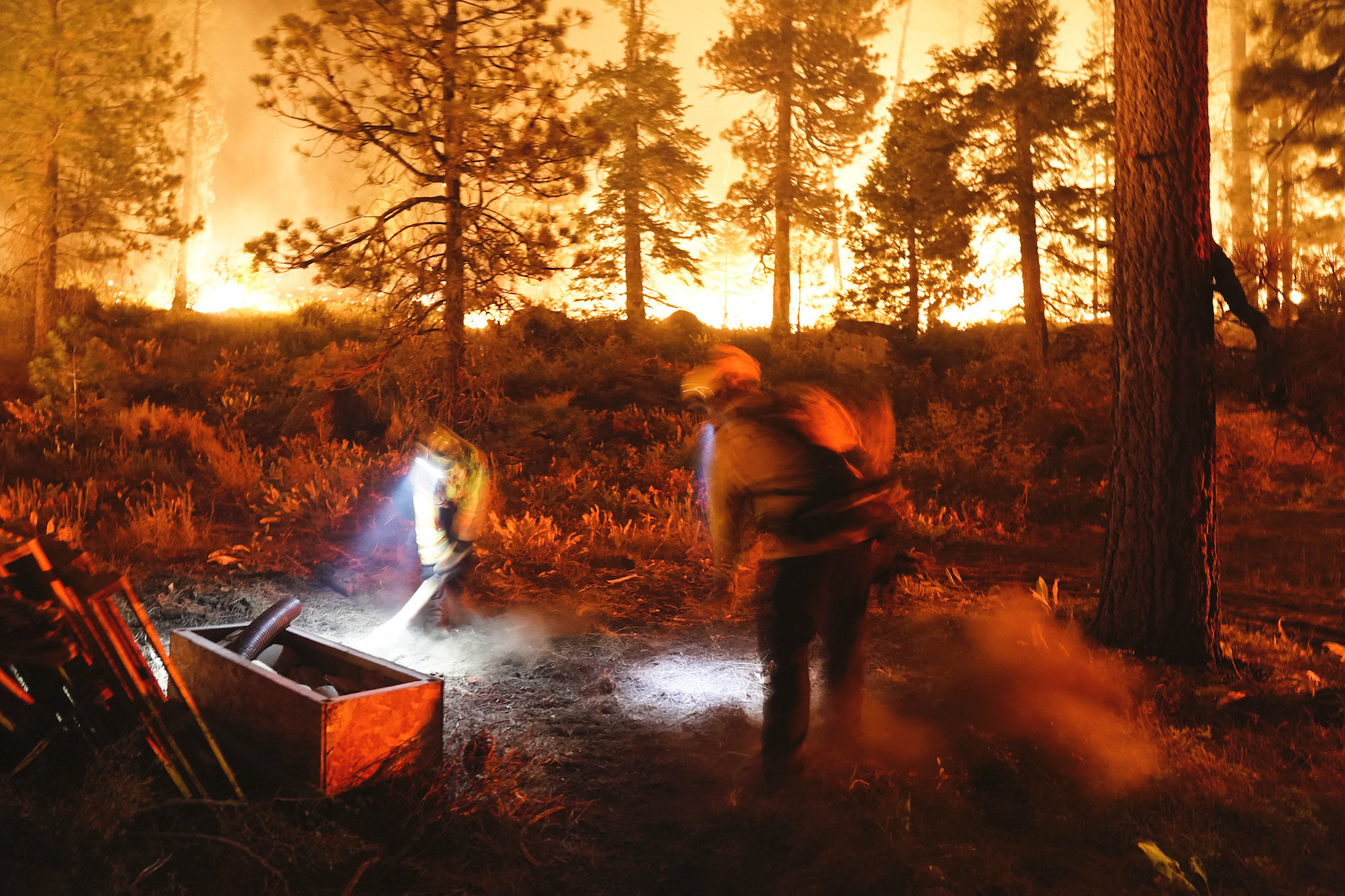 Two firefighters wearing gear and headlamps work at night in a burning forest