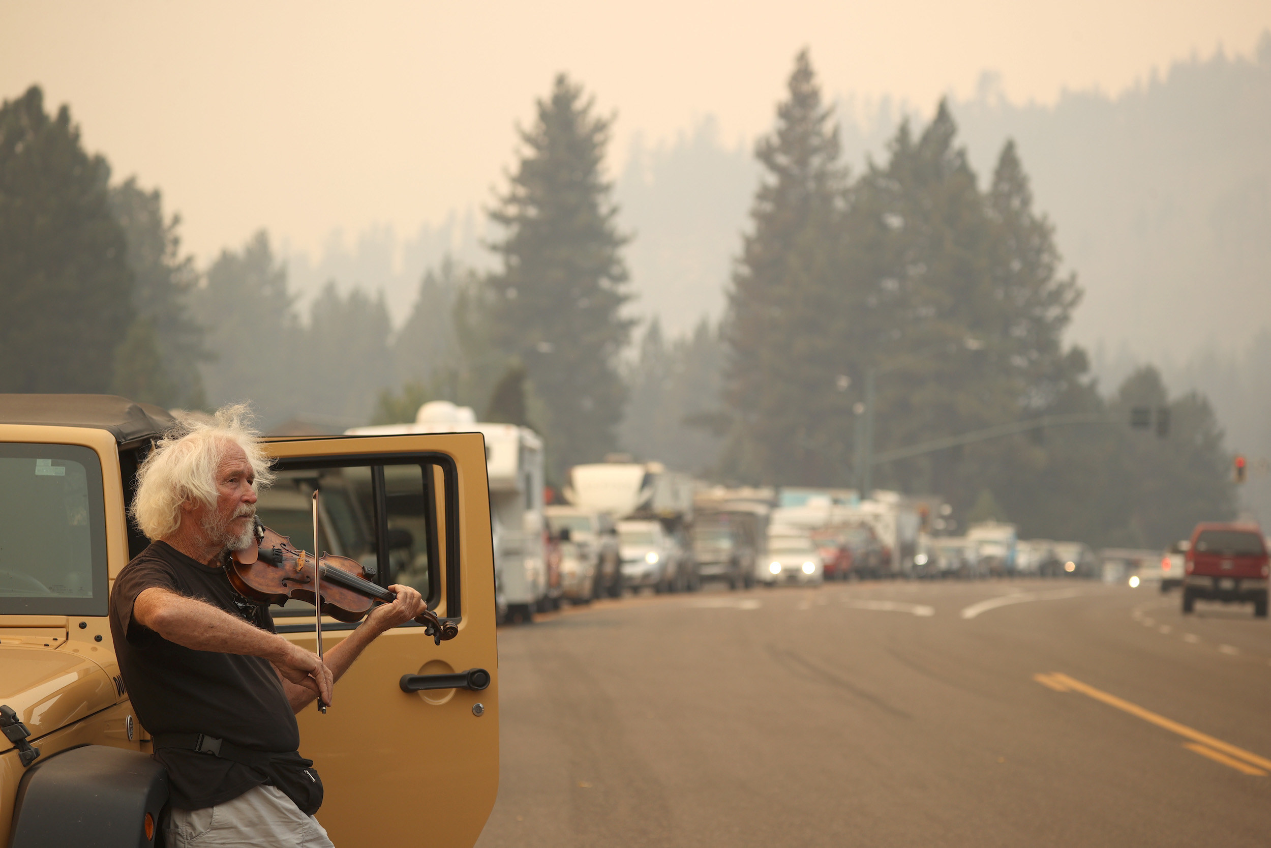 A man plays violin and leans against his Jeep in front of several cars in bumper-to-bumper traffic under a smoky sky