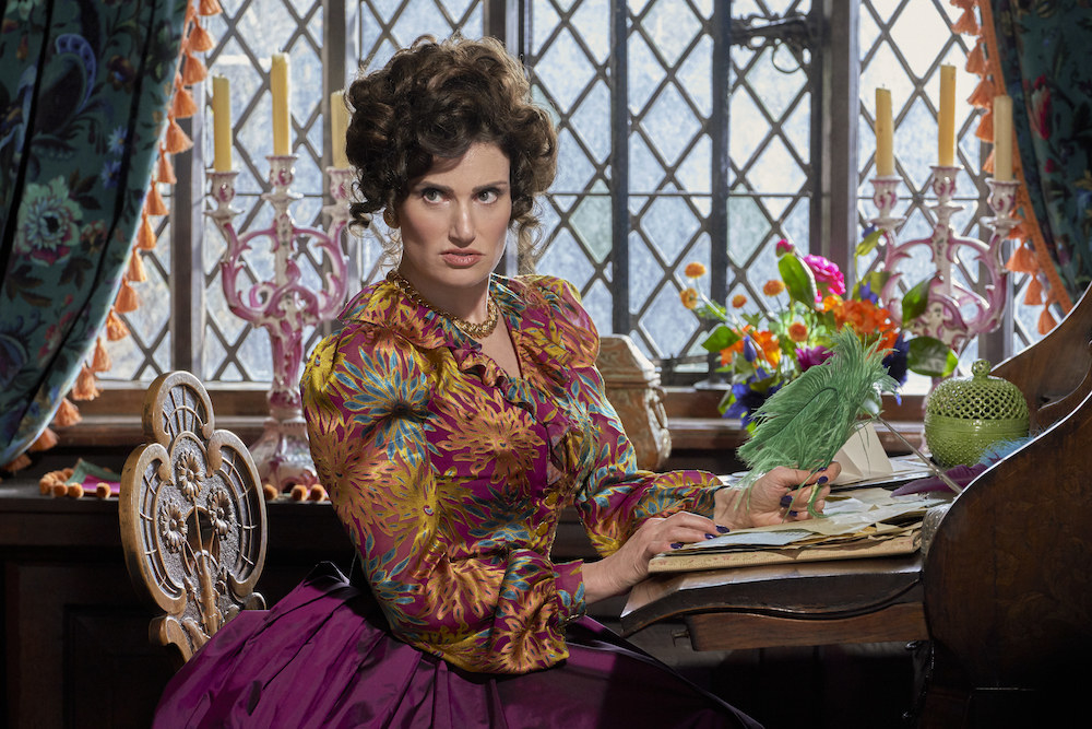 Idina as the stepmother, wearing a flower-decorated gown, sitting at a table writing letters