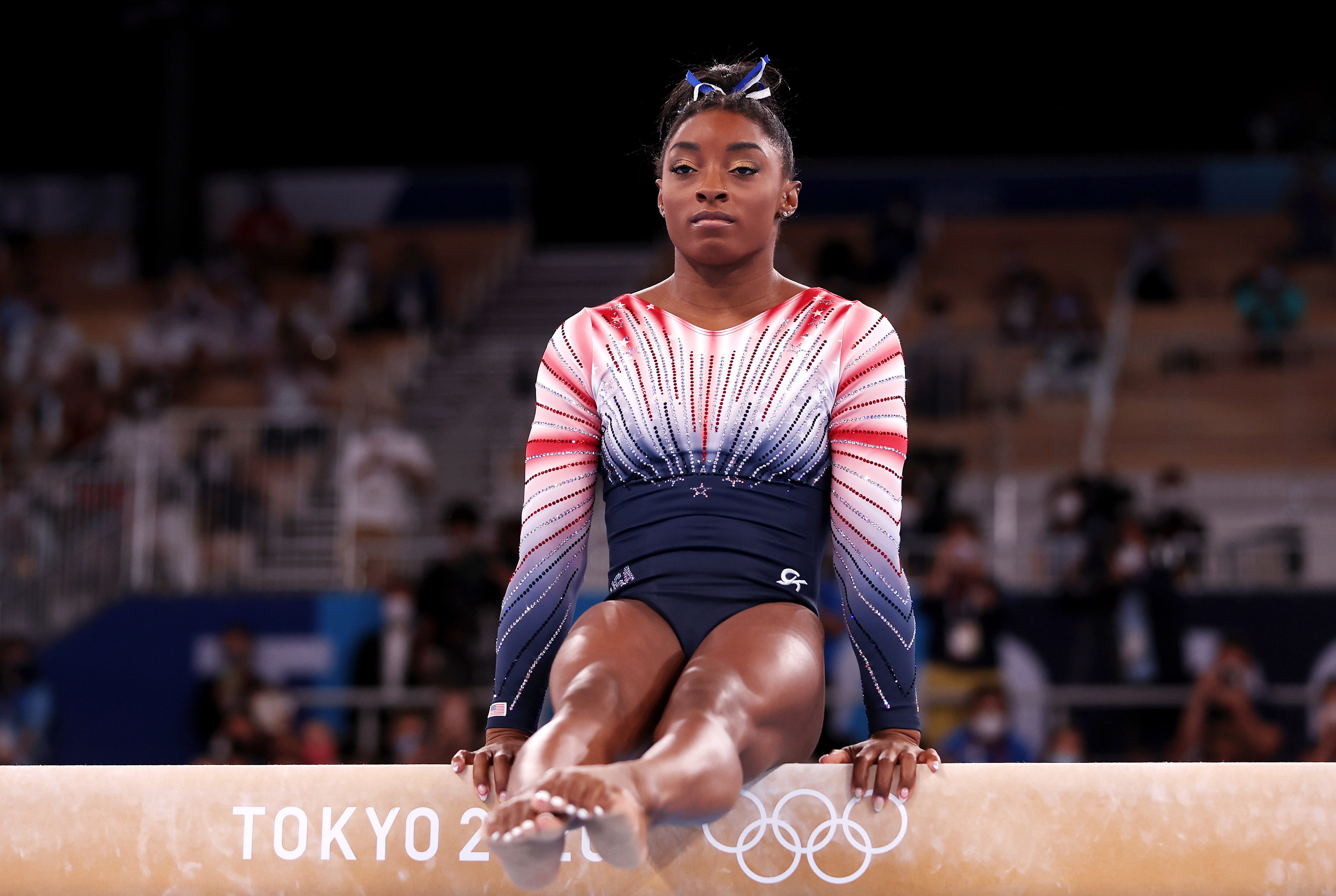 Biles in a seated position on the balance beam with her hands on the beam