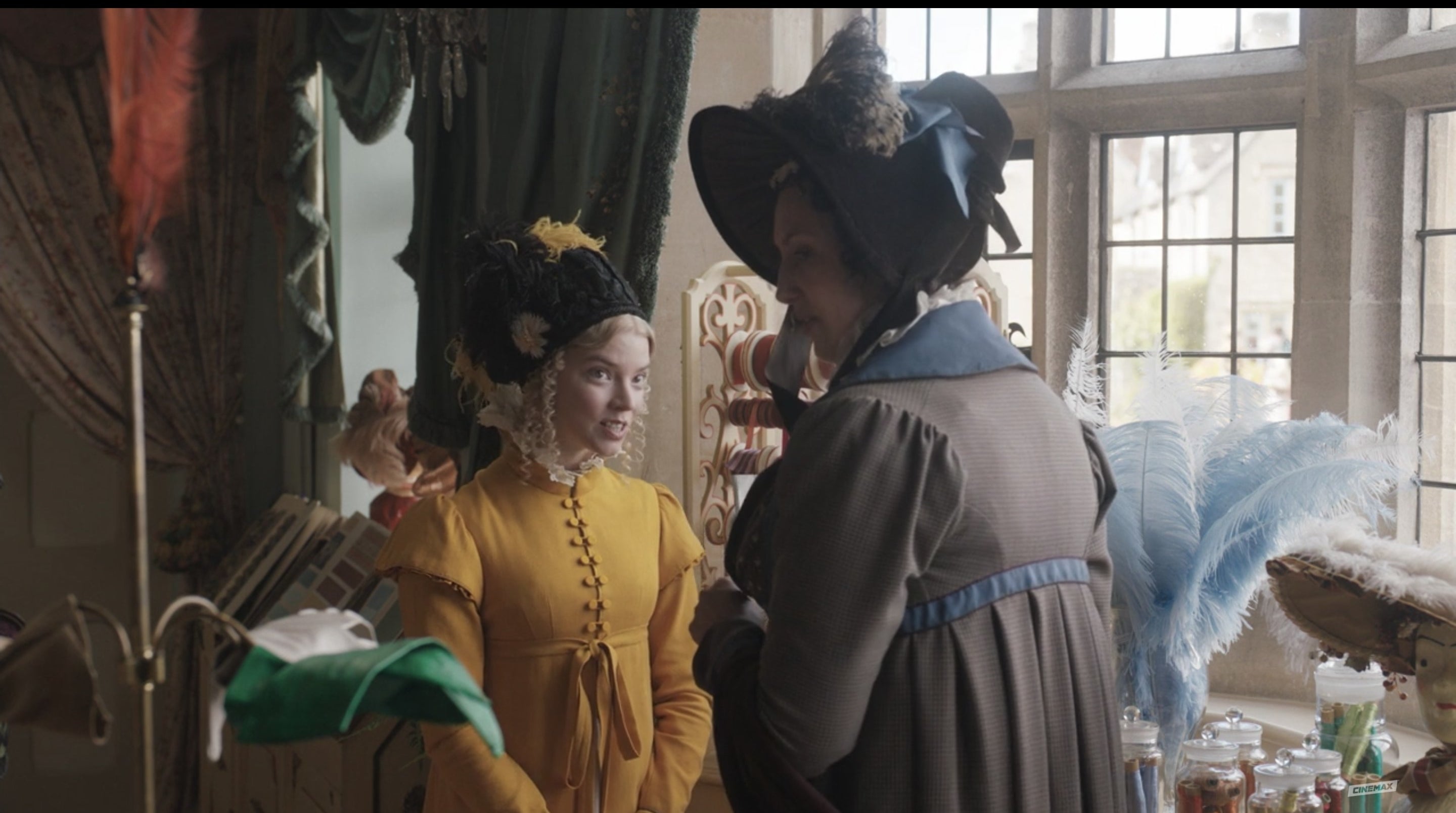 Emma wears a bright yellow dress and talking to a very distracted Miss Bates
