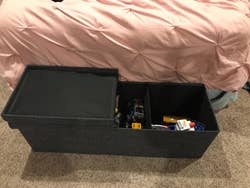 Reviewer's photo showing the dark grey ottoman at the foot of their bed with the lid open to reveal toys