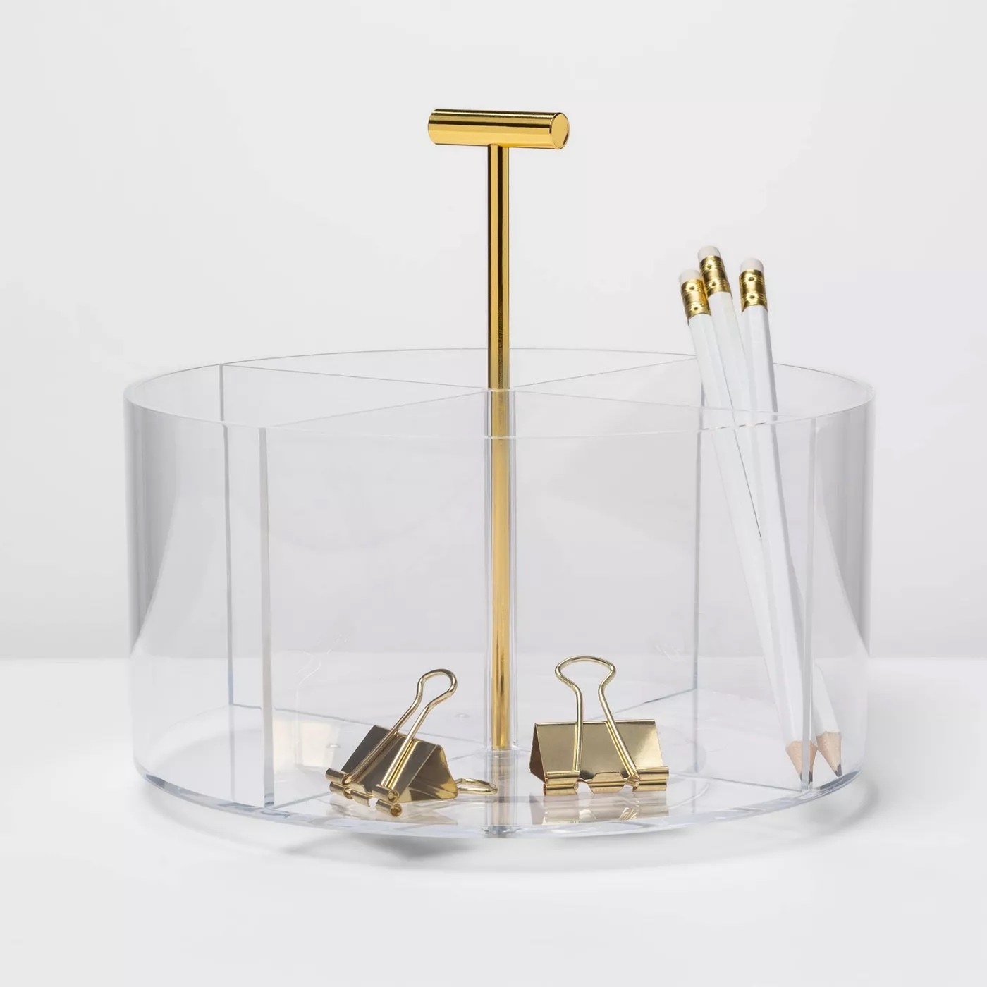 A clear round desktop organizer with gold and white pencils and gold paperclips.
