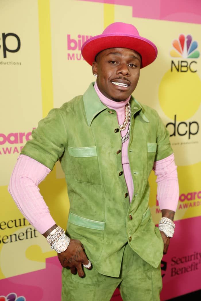DaBaby smiles for the camera while wearing a hat, suit, and sweater with multiple pieces of jewelry on both wrists