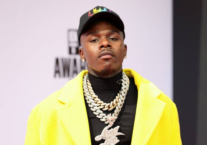 DaBaby wears a huge chain and a blazer and shirt with a hat as he poses for a picture
